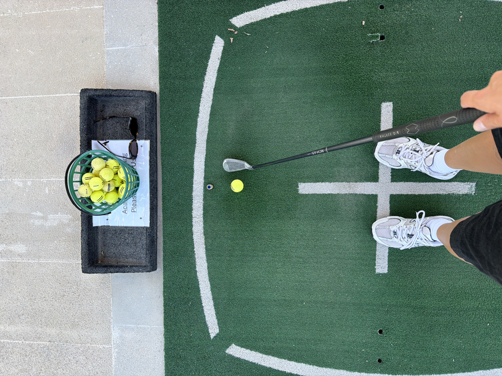 An overhead shot shows a pair of white athletic shoes standing on a green turf golf practice mat. The person wearing the shoes is holding a black golf club with a silver clubhead. Two yellow golf balls and a small blue object are on the mat in front of the person. A green basket filled with yellow golf balls sits on a grey mat to the upper left. A pair of dark sunglasses rests next to the basket. The practice mat is bordered by a white line and a light-colored tiled area. This scene is at a golf driving range in Tsada, Cyprus.