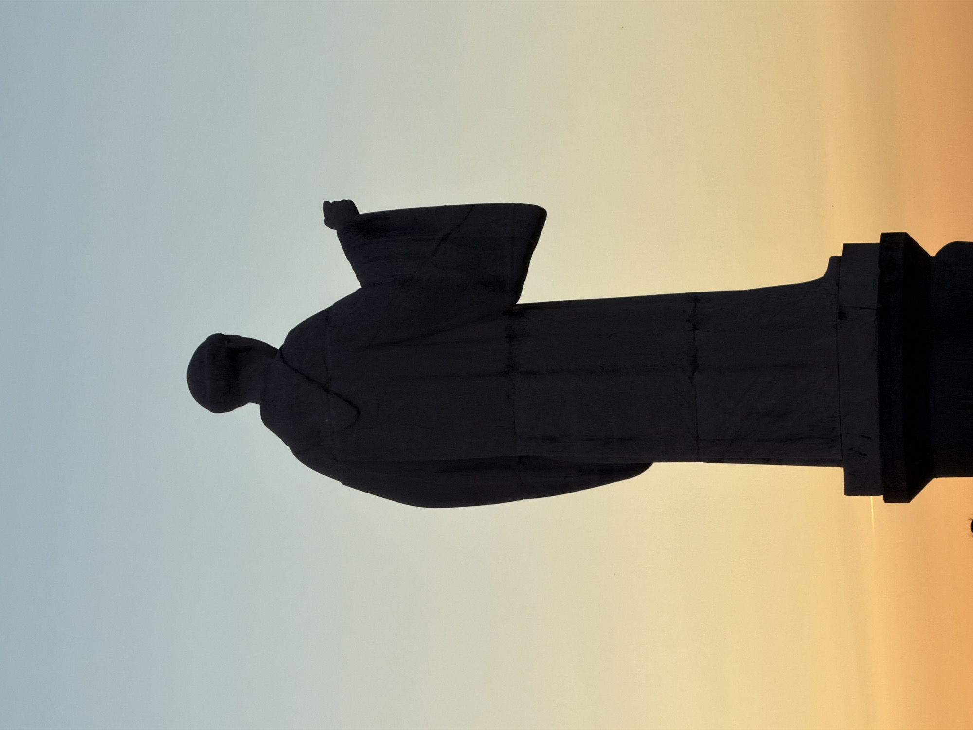 A silhouette of a statue stands against a gradient sky, transitioning from light blue to orange, likely during sunrise or sunset. The statue is of a robed figure with a raised fist, mounted on a tiered pedestal. The statue is located in Bragança, Portugal.