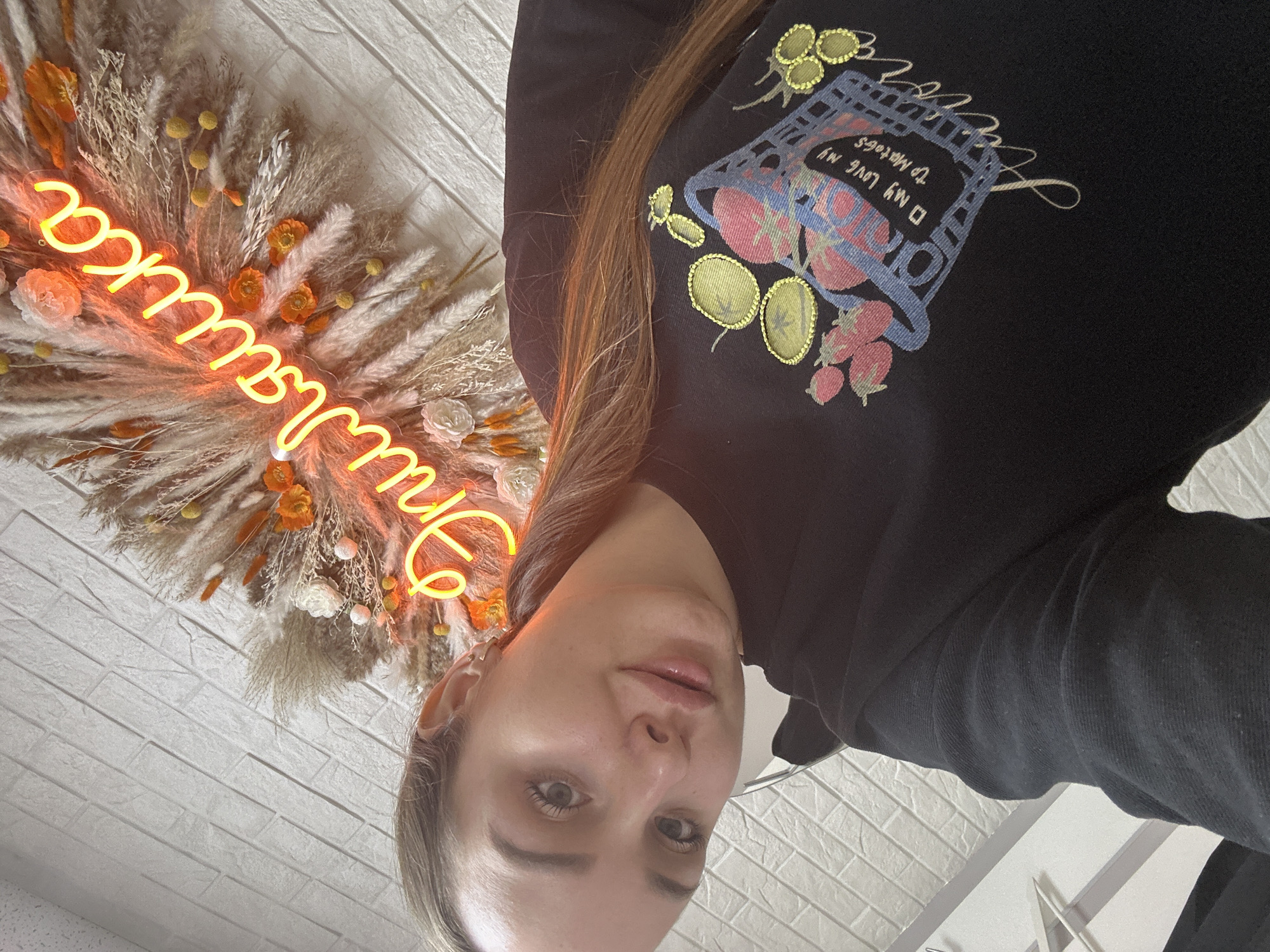 A woman is taking a selfie. She has light skin, light eyes, and long, light brown hair. She is wearing a black sweatshirt with a colorful design on the front. Behind her is a decorative wreath made of dried grasses and flowers, with an orange neon sign in the middle that reads "Natalia". The background is a white brick wall. The photo may have been taken in Chaikovsky, Russia.