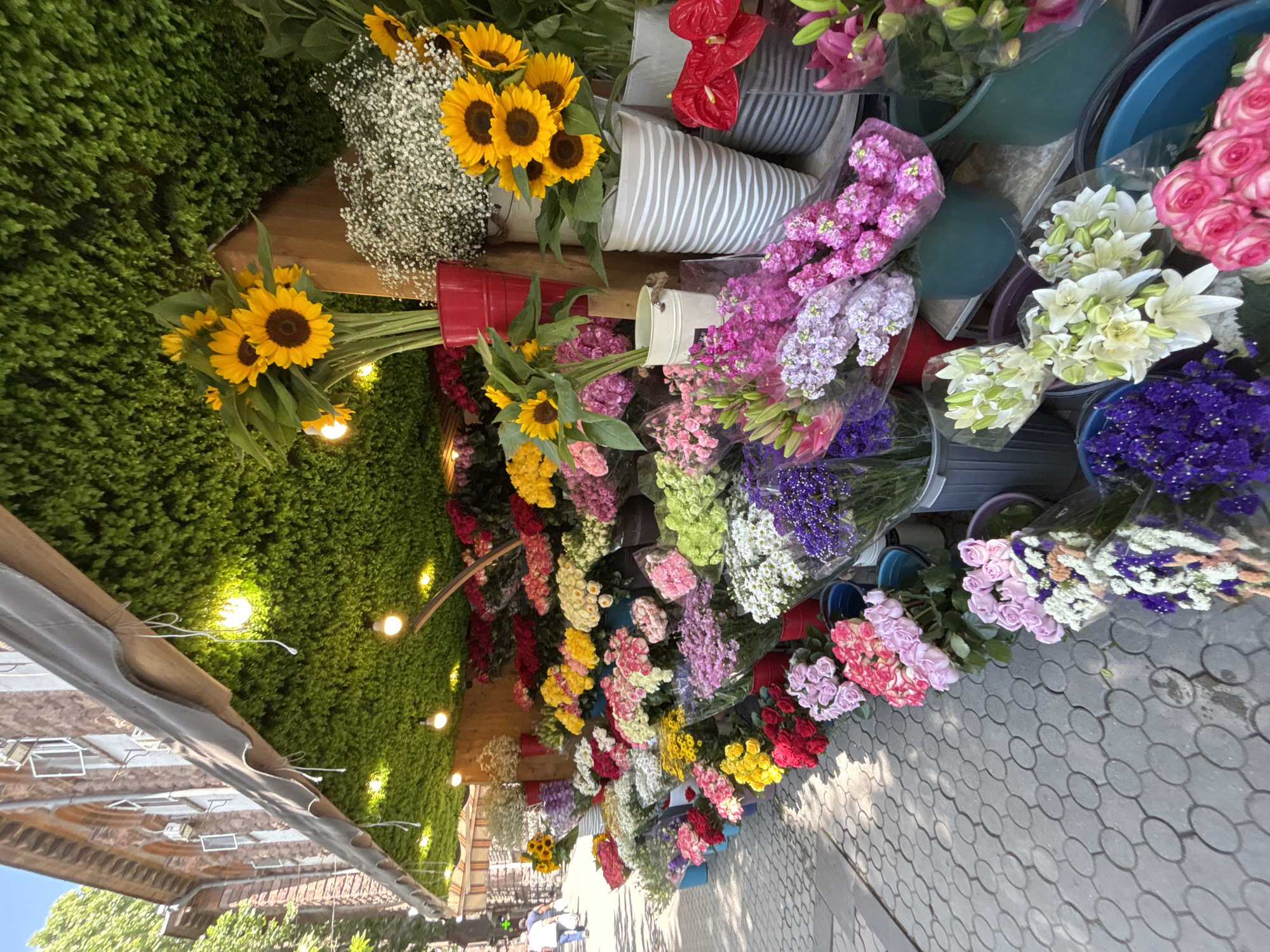 A street-level view captures a vibrant flower stall set up along a paved sidewalk in Yerevan, Armenia. The stall is covered by a green, leafy canopy with exposed light bulbs illuminating the flowers below. A red awning extends over the canopy, attached to a brick building in the background.
The stall is densely packed with a wide variety of colorful flowers displayed in buckets and vases. Prominently featured are bouquets of bright yellow sunflowers, pink and red roses, purple statice, white baby's breath, and various other mixed blooms. The flowers are arranged in layers, with taller arrangements at the back and shorter ones at the front.
In the background, a few pedestrians are visible walking down the sidewalk, one person appears to be a male in a white shirt and blue pants. The street is paved with cobblestones or interlocking pavers. The overall scene depicts a lively and colorful outdoor market in an urban setting.