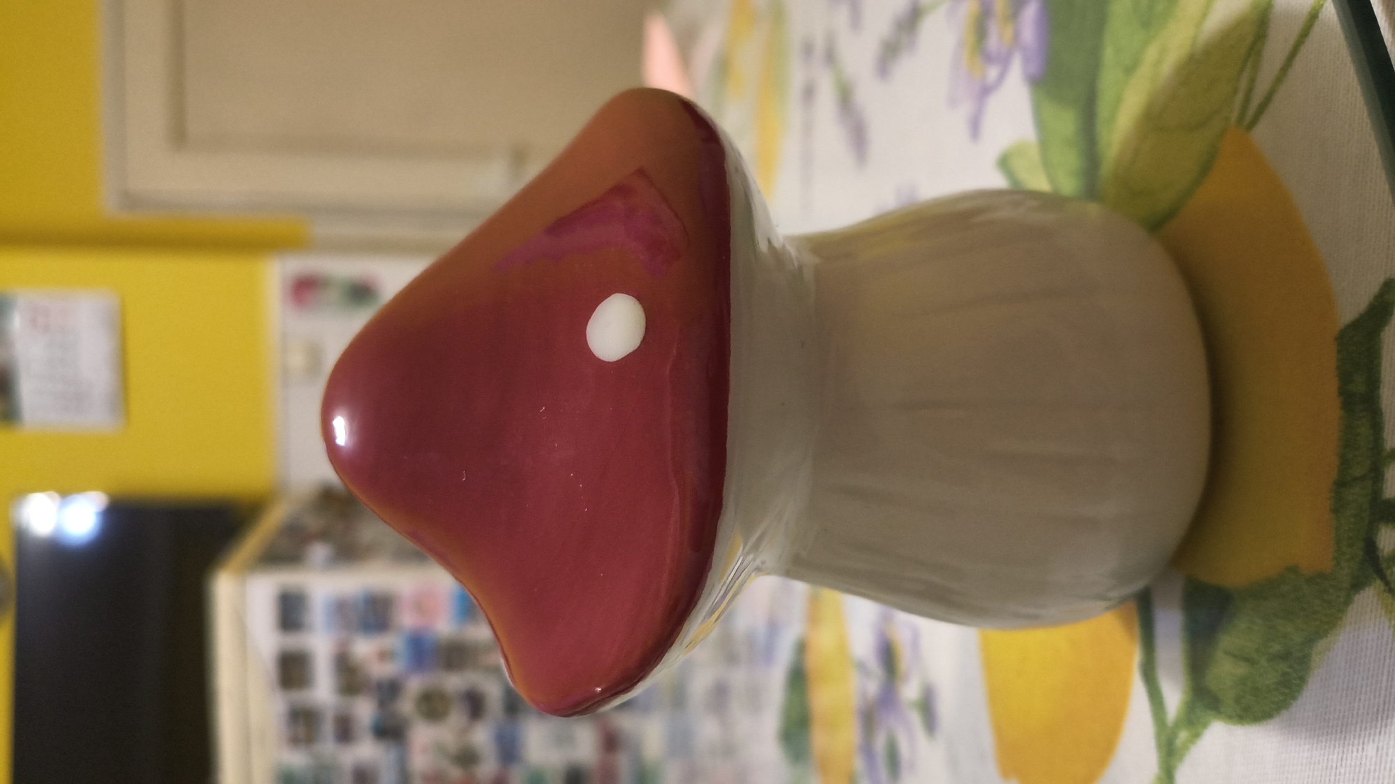 A glazed ceramic mushroom-shaped decorative object is centered in the frame, positioned on a patterned tablecloth. The mushroom features a highly reflective, domed cap in a deep reddish-burgundy hue, with a single prominent white circular spot located on the upper left side of the cap. Light reflections are visible on the glossy surface of the cap. The stem of the mushroom is off-white or light beige, with a matte finish and subtle vertical texturing.
The tablecloth beneath the object is white with a repeating design incorporating yellow lemon slices or whole lemons with green leaves and stems, alongside clusters of small purple floral elements.
The background is significantly out of focus, indicating shallow depth of field. A bright yellow wall dominates the mid-ground. On the left side, blurred dark shapes, possibly a television or monitor, show bright, undefined light sources reflecting from their surface. Below this, a lighter-toned shelving unit or cabinet displays numerous small, indistinct rectangular items. On the right side, a light-colored doorframe and a partially visible door are present against the yellow wall. A white rectangular object, resembling a calendar, is affixed to the yellow wall in the upper-center background. The scene depicts an indoor setting, likely a domestic environment, located in the city of Shumen, Bulgaria.