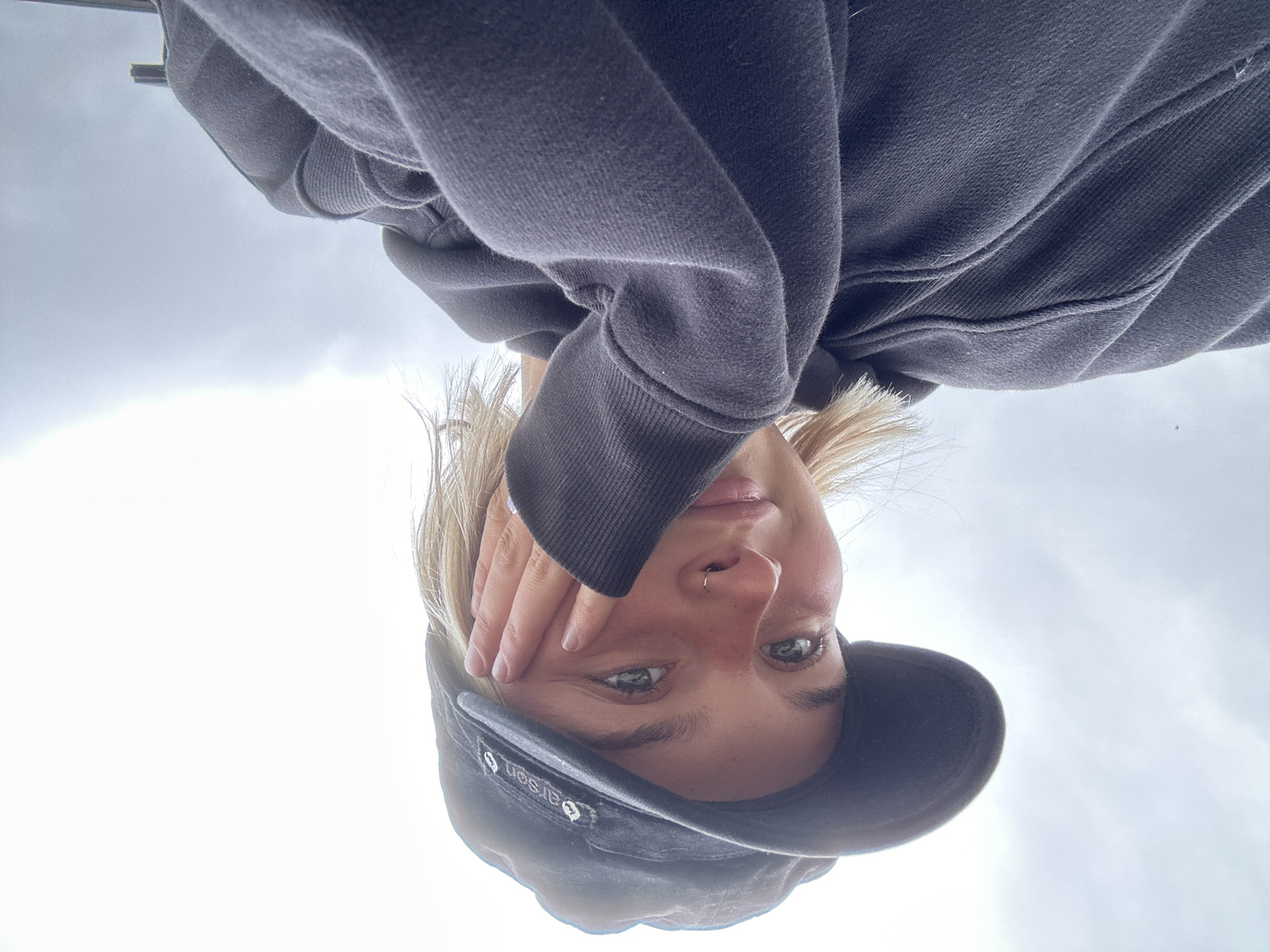 A medium shot captures a young woman from the chest up, looking up and to her left with her left hand resting on her cheek. She is wearing a dark grey baseball cap and a dark grey hoodie. The background is a bright, overcast sky. The image appears to have been taken outdoors, possibly in Whistler, Canada, given the cloudy sky and the casual attire.