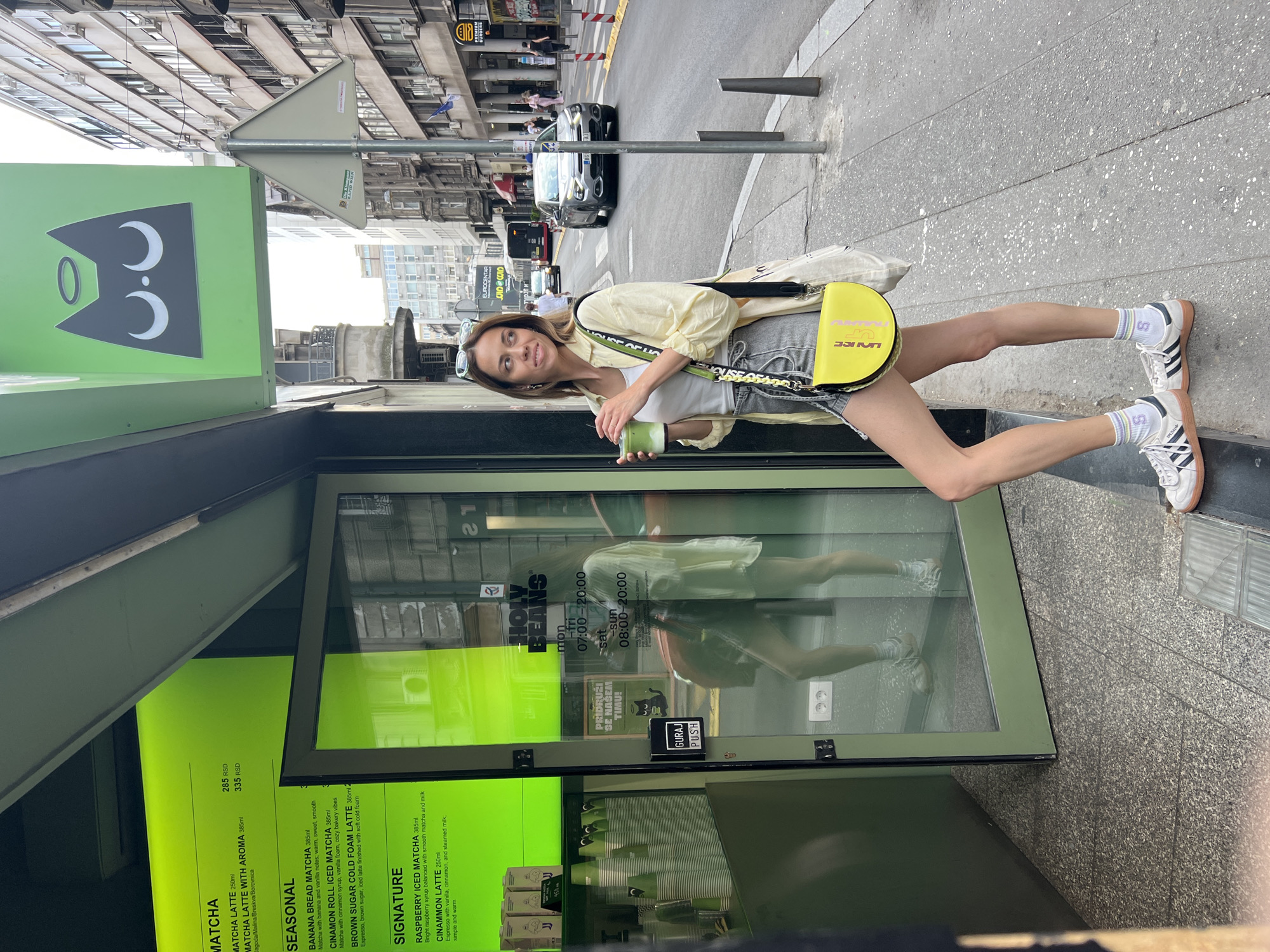 A young woman stands on a sidewalk next to the "Holy Beans" coffee shop in Belgrade, Serbia, holding a green beverage. She is wearing a light yellow shirt, grey denim shorts, a white top, white sneakers with black stripes, and a yellow purse with a strap that has green and black lettering. The coffee shop has a green exterior with a menu displaying "MATCHA," "SEASONAL," and "SIGNATURE" drinks in Serbian dinars (RSD). The shop's glass door reflects her image. The street scene includes passing cars and buildings, and a street sign is visible above the shop.