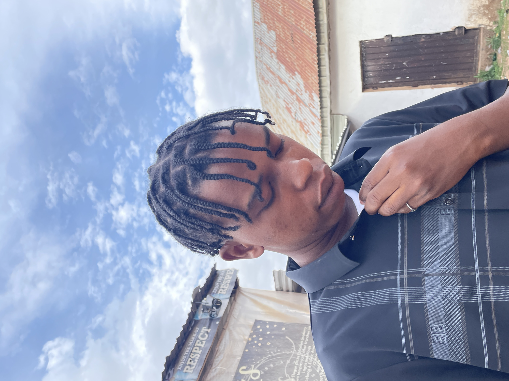 The image captures a young person with dark skin and dark, intricately braided hair, in a daytime outdoor setting. The individual is positioned slightly to the right of the frame, facing generally left but looking downwards with their eyes mostly closed or cast away. They are wearing a dark, possibly navy blue or black, collared shirt with a distinct plaid pattern in lighter grey and white lines. A white t-shirt is visible underneath the collar. The person's right hand is raised to their neck, seemingly adjusting the collar or buttoning the shirt. A ring is visible on a finger of their right hand.
The background features a bright, partly cloudy blue sky, prominently displayed in the upper left. Behind the person, to the upper right, is a light-colored building with a dark, possibly wooden, framed window or door, and a weathered corrugated metal roof. In the lower left, another structure or stall is visible, bearing various signs. Text deciphered from these signs includes phrases like "ENJOY THE MARK OF RESPECT," "COLD RESPE," and mentions of locations or entities such as "Elegun plaza Entinders academy," "Lagas and Abuja," and "cebook & fiktek:". The shirt also displays a logo that includes the letters "BB," stylized as part of the plaid pattern. The overall atmosphere suggests a casual, possibly urban or semi-urban environment in Jos, Nigeria.