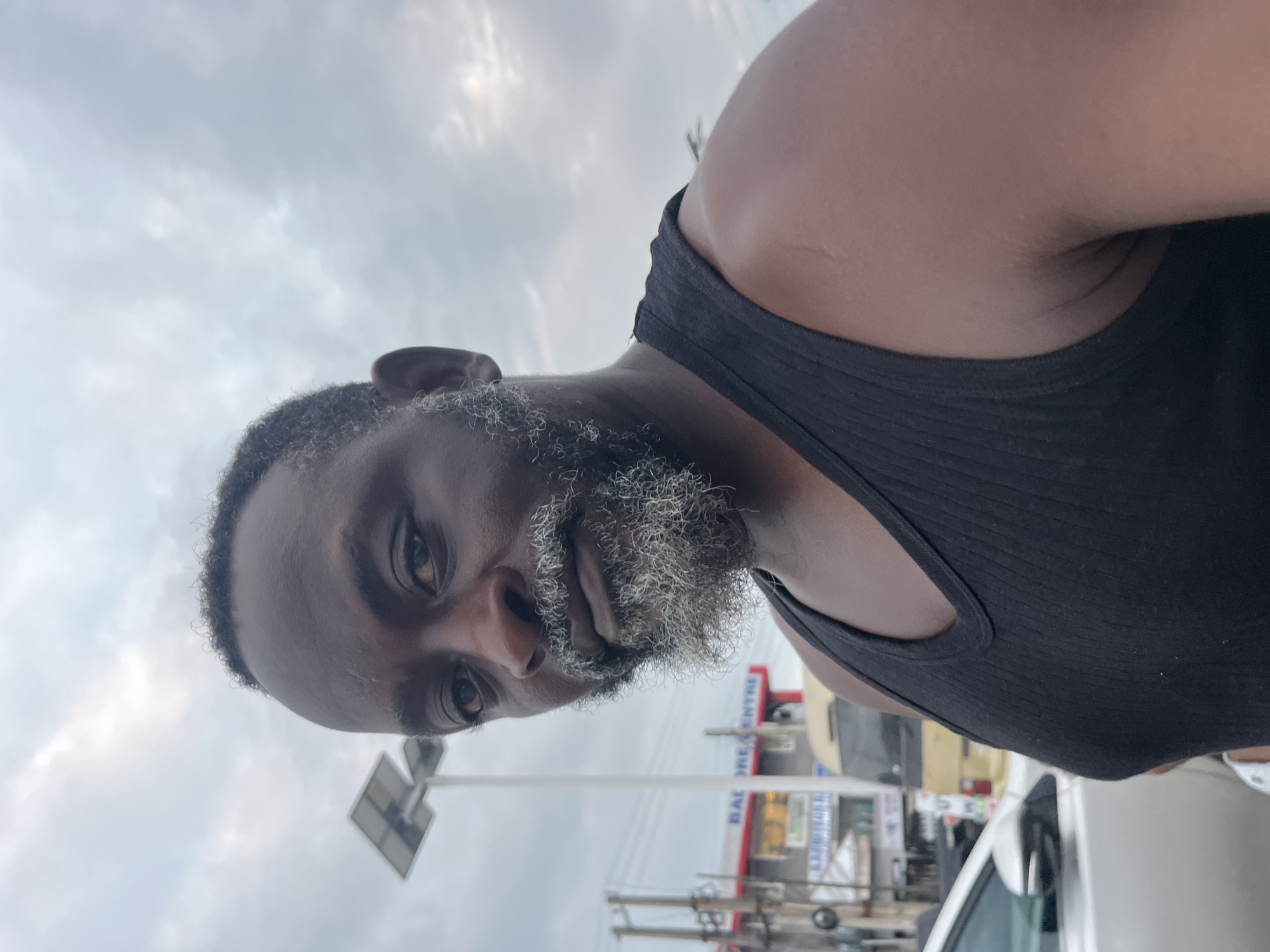 A close-up, eye-level selfie shows a Black man with a graying beard and a black tank top. He is in what appears to be a city street setting. In the background, there is a street light with solar panels, buildings with signage, and vehicles. The sky is overcast. One of the buildings in the background has a sign that reads "BALORE CENTRE". This indicates the location is Ado, Nigeria.