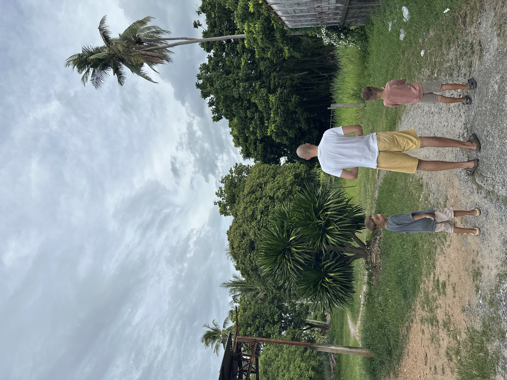 In Baan Mae Nam, Thailand, a man with a shaved head, wearing a white t-shirt and yellow shorts, stands facing away from the camera, looking towards a path. To his left and slightly in front, a young boy in a gray t-shirt and beige shorts also faces away. To the man's right and in front, another young boy in a pink t-shirt and gray shorts faces away. They are on a dirt and gravel path surrounded by lush green vegetation, including trees and palm trees. The sky is overcast with gray clouds, and a dilapidated wooden structure with a corrugated metal roof is visible to the far left. To the right, a weathered wooden building with graffiti is partially visible. The scene appears to be a rural or semi-rural setting.
