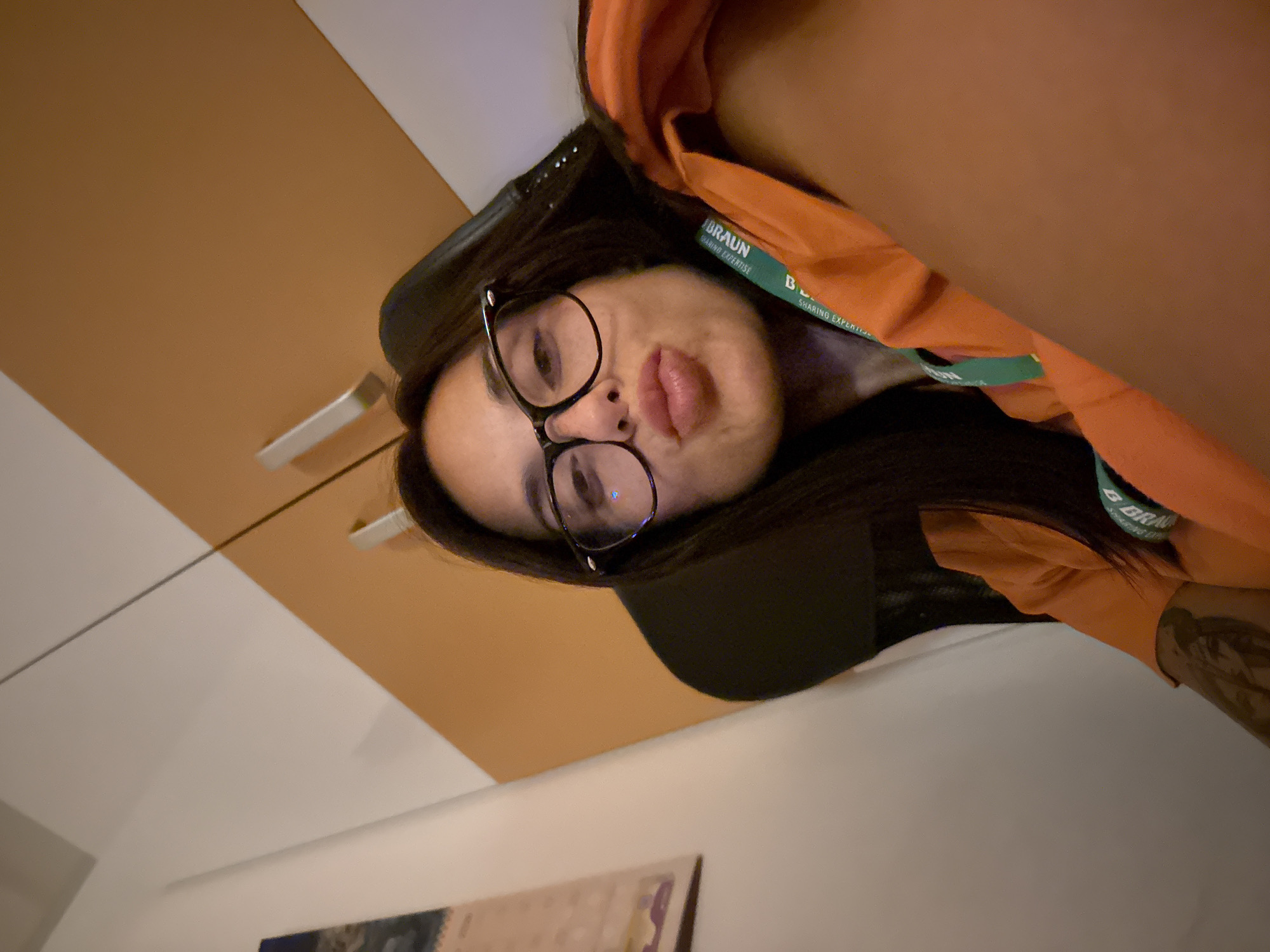 A woman with dark hair and glasses is taking a selfie in a room. She is wearing an orange shirt and a green lanyard with text on it that is partially obscured. The background shows a wall with two cabinet doors. A calendar is visible on the left side of the frame.
