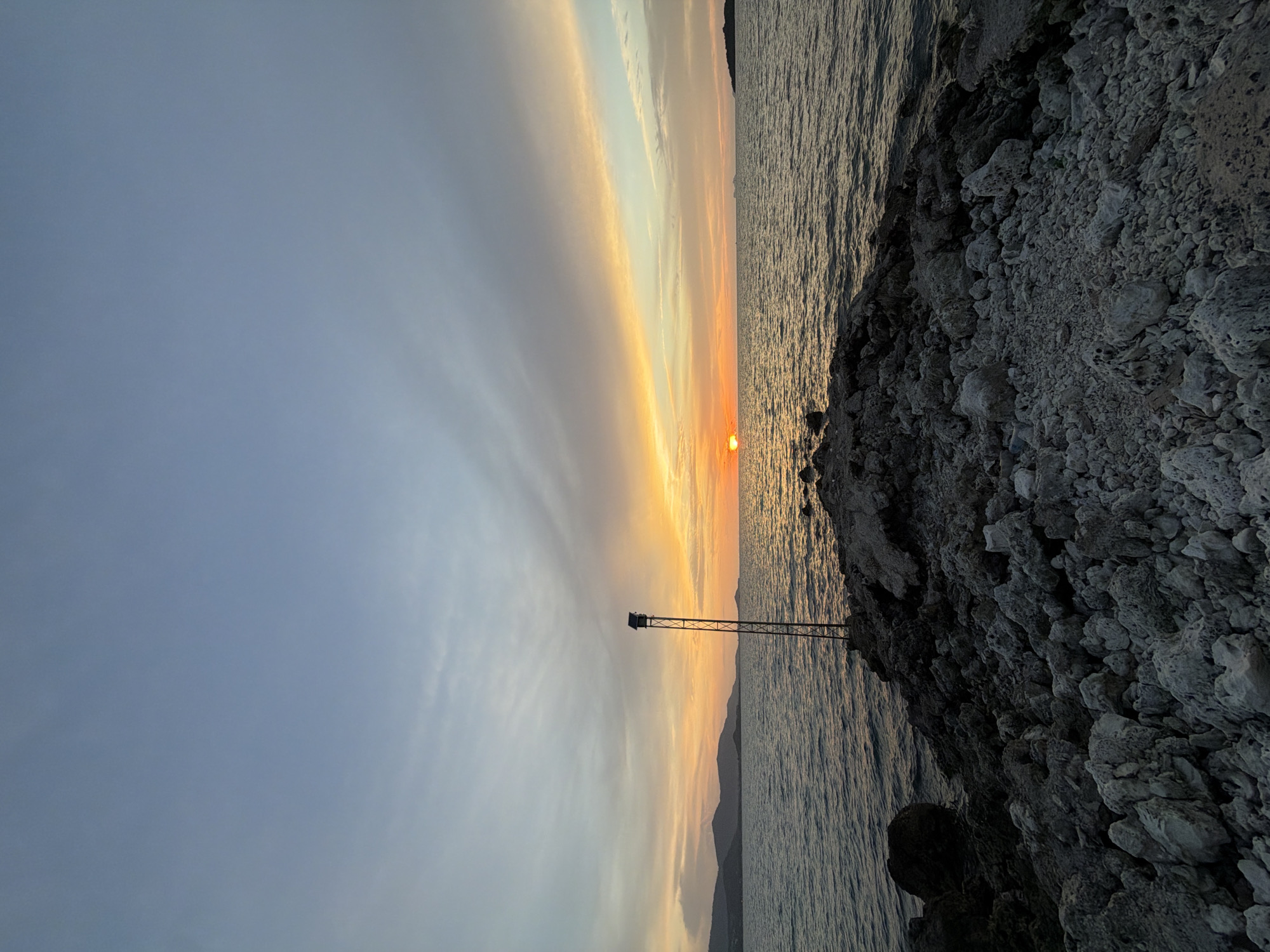 A wide-angle, eye-level shot captures a tranquil sunset over the Gulf of Thailand, likely from the rocky shores of Ko Samui District, Thailand. The sun, a small orange orb, is setting on the horizon, casting a warm glow across the water and sky. Wispy clouds streak across the pale blue sky, tinged with orange and yellow from the setting sun. The sea is calm, with gentle ripples reflecting the colors of the sky. In the foreground, a rocky, uneven shoreline extends into the water. A tall, slender metal pole with a small box-like structure at its top stands on the rocks, positioned to the left of center. In the distance, a dark, silhouetted mountain range borders the horizon on the left, and a small island is visible to the right. The scene is devoid of people or any discernible activity, presenting a serene and natural landscape.