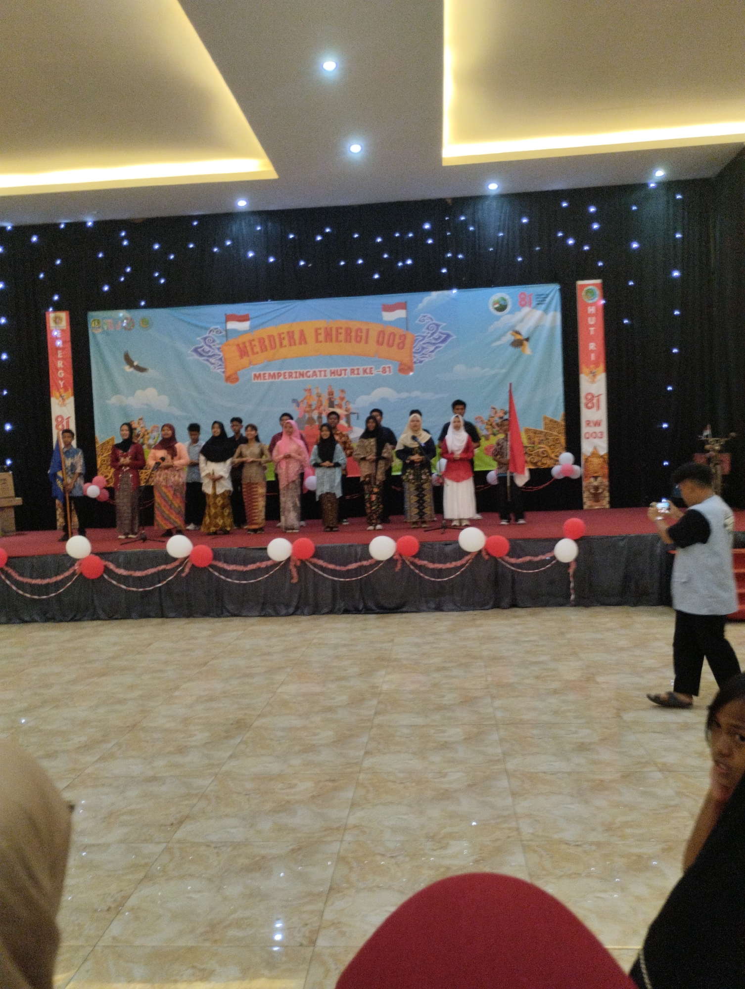 This is a photograph of a stage performance at a formal event, likely a celebration or competition. The stage is decorated with a large banner that reads "MERDEKA ENERGI 003 MEMPERINGATI HUT RI KE - 81". This banner indicates the event is related to Indonesian Independence Day (HUT RI) and is possibly sponsored by an energy company.
On the stage, a group of approximately 12-15 individuals, mostly young women, are lined up, some holding microphones as if about to perform or having just finished. They are dressed in traditional Indonesian attire, including batik skirts and colorful tops, some with headscarves. In the background, there's a black curtain adorned with small white lights, creating a starry effect. Red and white balloons are placed along the front edge of the stage.
To the right of the stage, a man in a light-colored vest and dark pants is seen holding up a smartphone, appearing to be filming or photographing the event. The flooring of the venue is tiled with a beige and brown marble pattern. The ceiling features modern, recessed lighting. The overall atmosphere suggests a community or school event.
While the image itself does not explicitly state the location in Bekasi, Indonesia, the context of "HUT RI" strongly suggests it is an event celebrating Indonesian Independence Day, a national holiday observed throughout the country, including in Bekasi. The banner's text "MERDEKA ENERGI 003" might refer to a specific event or competition related to energy and independence within that region.