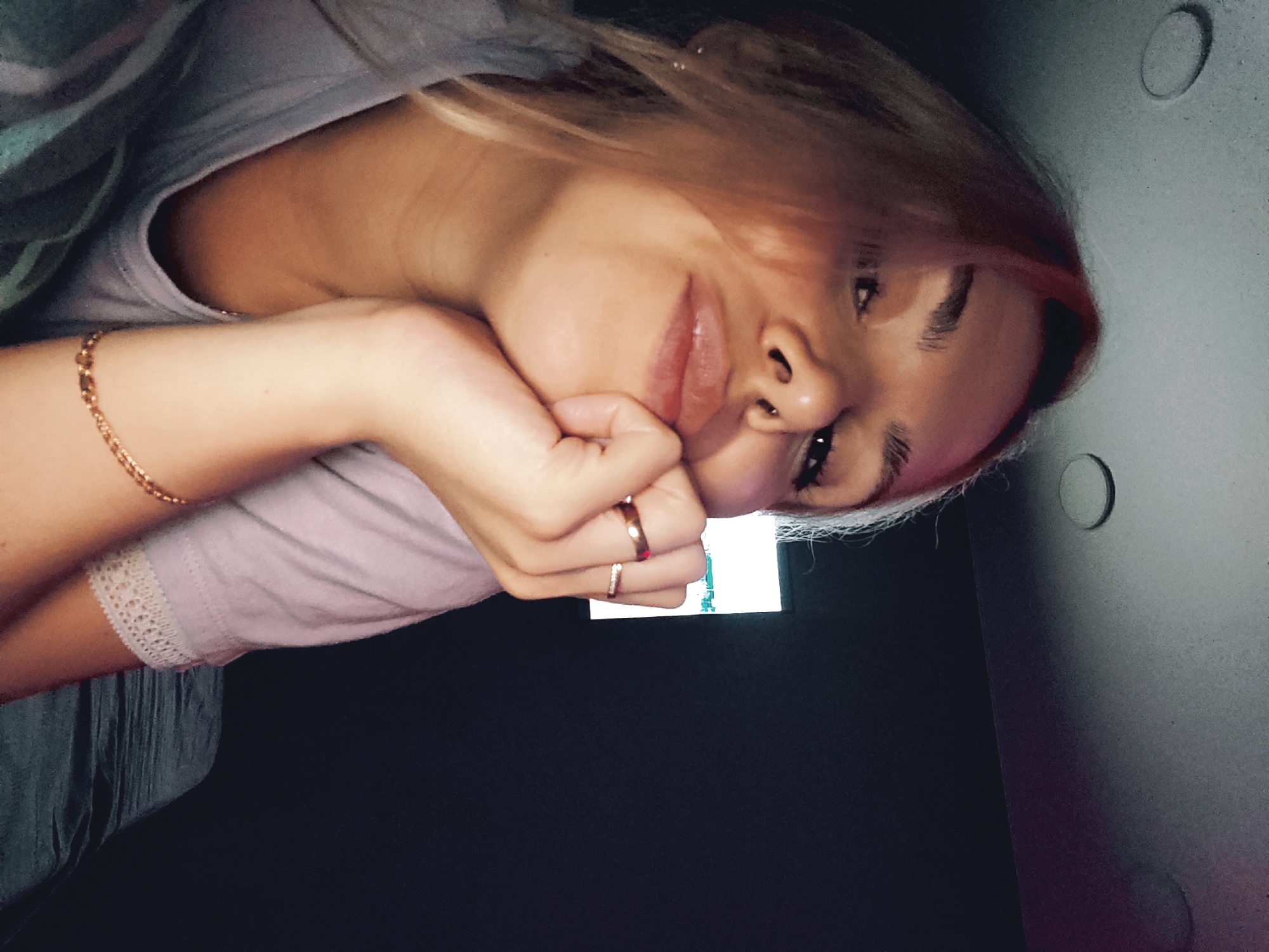 A young woman is captured in a close-up, rotated portrait, looking towards the viewer with a gentle smile. Her head is tilted, resting on her right hand, which is adorned with a thin gold-colored chain bracelet and at least two rings. She has light-colored hair with reddish or pinkish streaks, and a small ring piercing in her left nostril. She wears a light-colored top, possibly lavender or light grey, with a delicate lace trim visible on the sleeve near her elbow. Another darker garment or fabric is visible around her upper body.
The setting appears to be indoors and dimly lit. In the background, there's a dark, unidentifiable surface, and under her right hand, a bright, rectangular light source is visible, likely a phone or tablet screen, showing some indistinct graphics. To the right, a lighter greyish-blue surface features two distinct circular indentations or fixtures. The overall mood conveyed by her expression is relaxed and content. The image provides no specific visual cues to its location in Chaikovsky, Russia, other than the provided context. No discernible text is visible.