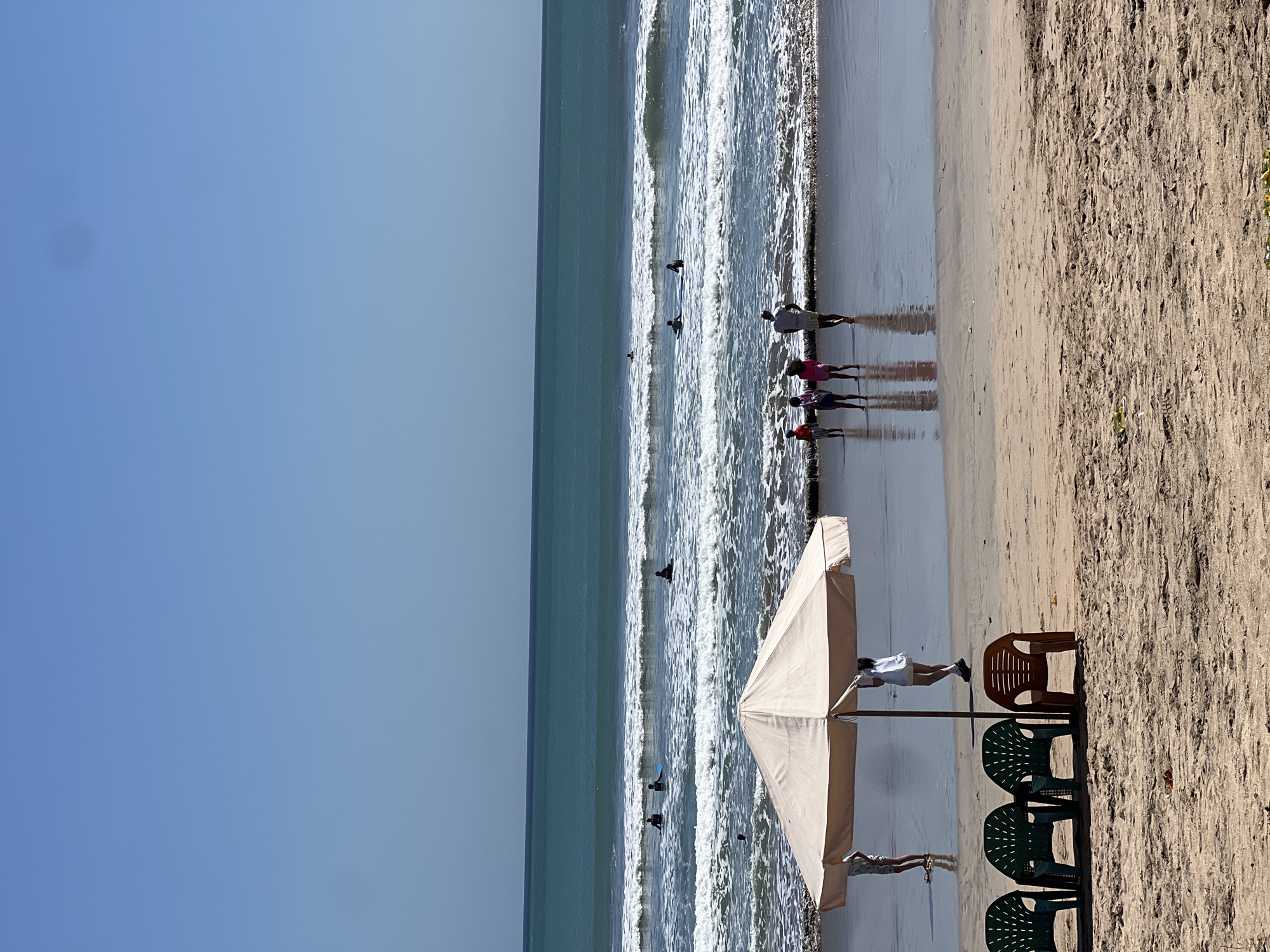 The image depicts a beach scene in Kuta, Indonesia, under a clear blue sky. In the foreground, four plastic chairs are arranged on the sand, three green and one orange. Behind the chairs, a large, beige beach umbrella shades two figures, one appearing to be a child and the other an adult. The sand is a light brown color with visible footprints and some small debris.
The middle ground shows the shoreline where gentle waves are breaking. Several surfers are visible in the water, either sitting on their boards or paddling. The ocean stretches to the horizon, with a distinct turquoise hue. Further back, a group of people, including adults and children, are standing on the wet sand near the water's edge. Some are facing the ocean, and one adult appears to be walking away from the water. The overall atmosphere is one of a relaxed beach day.