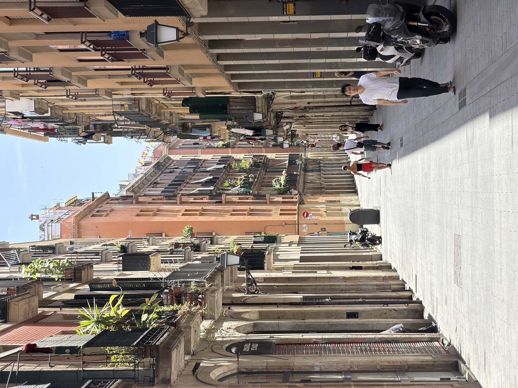 This is a street-level, medium shot of a pedestrian street in Barcelona, Spain, likely in the Gothic Quarter or El Born district, identifiable by the narrowness of the street, the architecture of the buildings with their balconies adorned with plants, and the general ambiance. The buildings lining the street are tall and multi-story, with facades in varying shades of orange, peach, and muted pink. Many balconies feature iron railings and are decorated with potted plants, some with vibrant green leaves. Laundry is seen drying on some of the balconies.
The street is paved with light-colored cobblestones, and the midday sun casts strong shadows from the buildings onto the walkway, creating a dappled light effect. Several people are walking along the street. In the foreground, a man wearing a white t-shirt and dark pants walks towards the camera, his face turned slightly to the side. Further down the street, a couple walks away from the camera; the woman is wearing a red skirt and a dark top, and the man is in a white t-shirt and shorts. Another woman in a white top and dark skirt walks in the same direction. In the background, a few more individuals are visible, their forms less distinct.
Several motorcycles and scooters are parked along the right side of the street. Street lamps with black metalwork are mounted on the walls of the buildings. The sky above is clear and blue. The overall impression is of a typical sunny day in a lively European city.