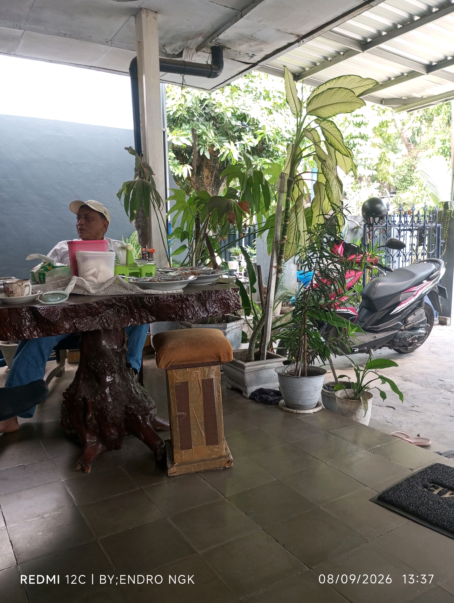 A middle-aged man wearing a white baseball cap and a white shirt sits at a large, rustic wooden table in an outdoor covered area, possibly a patio or veranda, in Nganjuk, Indonesia. The table is laden with plates, bowls, a pink container, and a tissue box. In front of him, on the table, are what appear to be snacks or food items, and a cup of coffee. A motorcycle, predominantly red and black, is parked to the right of the frame, with a black helmet resting on a metal fence behind it. Several potted plants, including a large one with broad leaves and a smaller, more slender one, are arranged around the seating area and the table. The floor is tiled with dark square tiles. In the background, lush green trees and foliage are visible, suggesting a tropical or subtropical environment. The overall scene depicts a relaxed, informal setting, possibly a home or a small eatery. The timestamp on the image indicates the date 08/09/2026 and time 13:37.