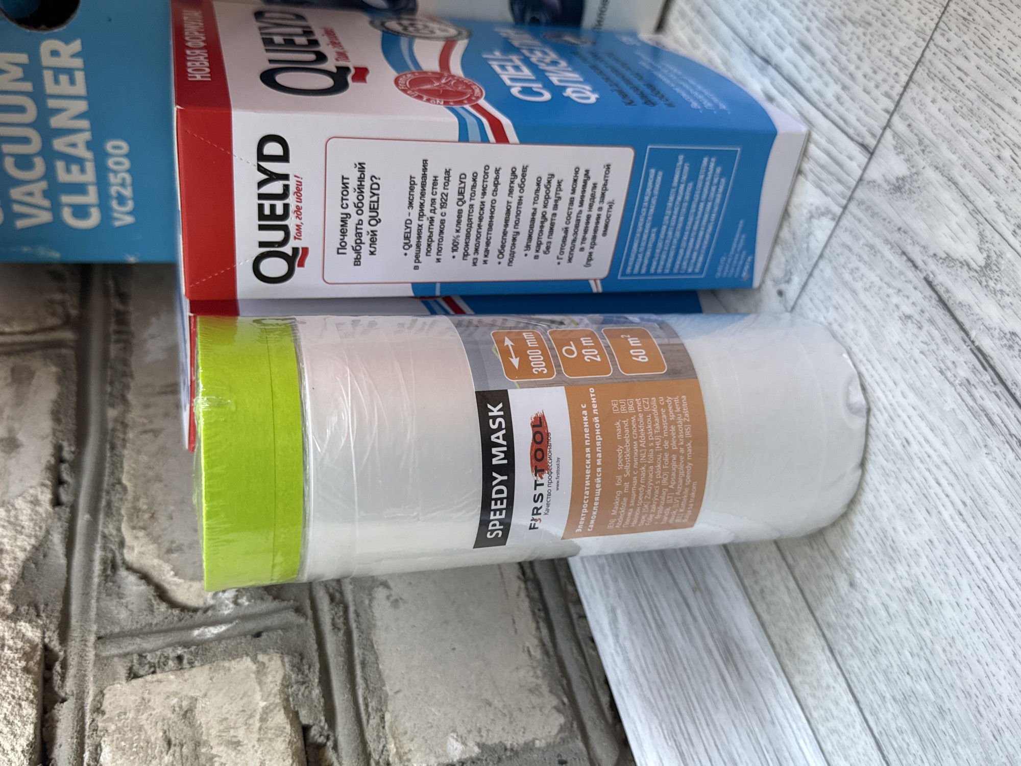 The image shows a close-up of two products on a wooden surface, with a textured concrete wall in the background. The product on the left is a roll of white masking film with a green top, labeled "SPEEDY MASK" and "FIRST TOOL" with a website address. The product on the right is a red and white box with the brand name "QUEL YD" prominently displayed, along with text in Russian. The box appears to be for wallpaper paste. There are also glimpses of other products in the background, including a blue box with "VACUUM CLEANER VC2500" printed on it, suggesting a retail or storage environment. The lighting is bright and appears to be natural, coming from above. There are no people or animals in the image, and no obvious signs of damage or destruction. The scene is static and appears to be a product display or inventory shot. The location is stated to be in Яромінскі сельскі Савет, Belarus.