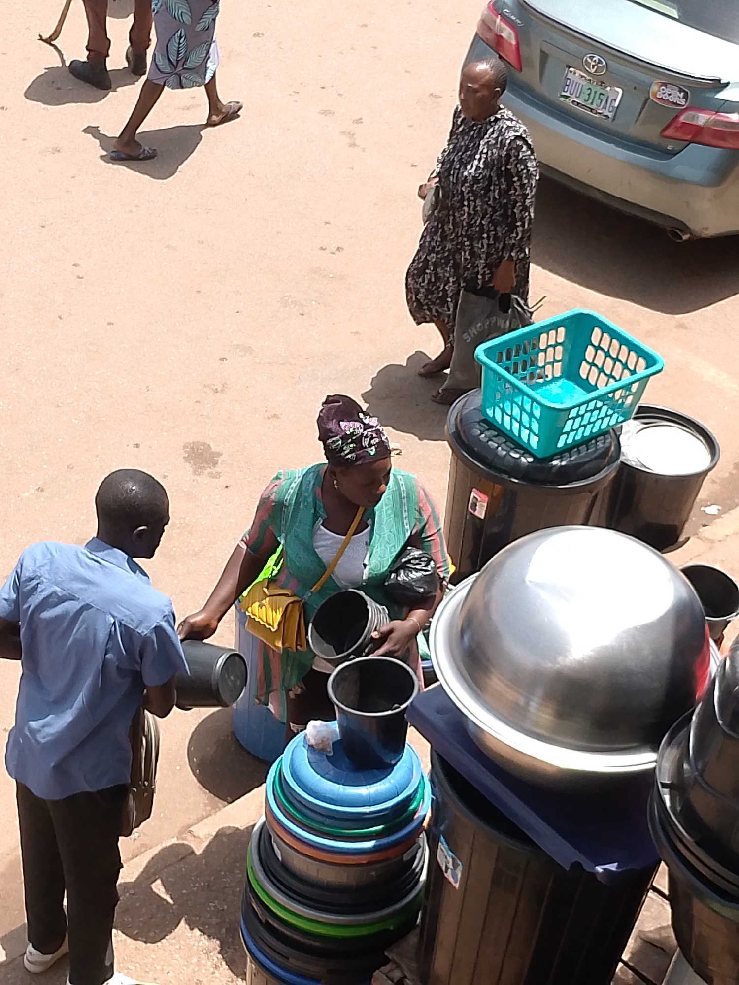 The image depicts an outdoor scene in Bukuru, Nigeria, with several individuals and a variety of objects, primarily containers, arranged on a sandy ground. In the foreground, a man wearing a blue shirt and dark pants is facing away from the viewer, holding a dark cylindrical object. To his right, a woman with a headscarf, wearing a green and pink striped garment, is interacting with the man and holding a yellow bag and a black bucket. Behind her, a large silver basin is prominently displayed on top of a black cylindrical container. Several other containers, including stacked colorful basins, black buckets, and a turquoise plastic basket, are visible throughout the scene. In the background, a silver car with a license plate reading "BUU 315AG" is parked. The overall environment suggests a market or roadside setting.