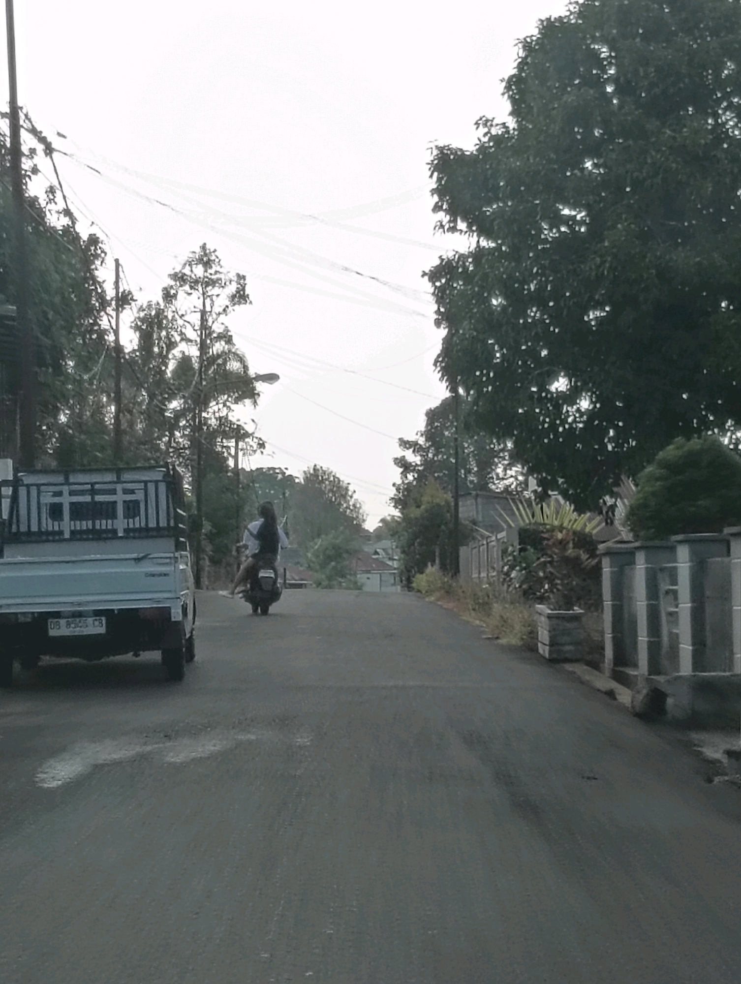 The image depicts a residential street in Tomohon, Indonesia, under an overcast sky. A light-colored utility truck is parked on the left side of the asphalt road, facing away from the viewer. Its license plate is partially visible. In the center of the road, a person is riding a dark-colored motorcycle, also facing away, wearing a white shirt and dark pants. The road is flanked by vegetation, trees, and houses with fences. A lamppost is visible further down the road. The overall scene suggests a quiet, possibly rural or suburban, setting.