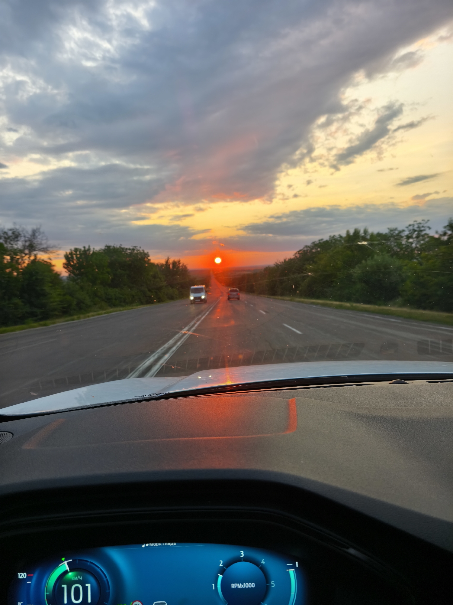 The image shows a perspective from inside a vehicle driving on a road towards a sunset. The sun is a bright orange orb on the horizon, casting a warm glow across the sky, which is filled with dramatic clouds. The road ahead is empty except for two vehicles, a white van and a smaller car, both illuminated by the setting sun. Lush green trees line both sides of the road, creating a natural backdrop. The dashboard of the car is visible in the foreground, showing a digital display with speed and RPM readings. There are no people visible outside the car, and the scene appears to be set in a rural or semi-rural area. The specific location in Cruglic, Moldova, is not explicitly indicated by any visual cues in the image.
