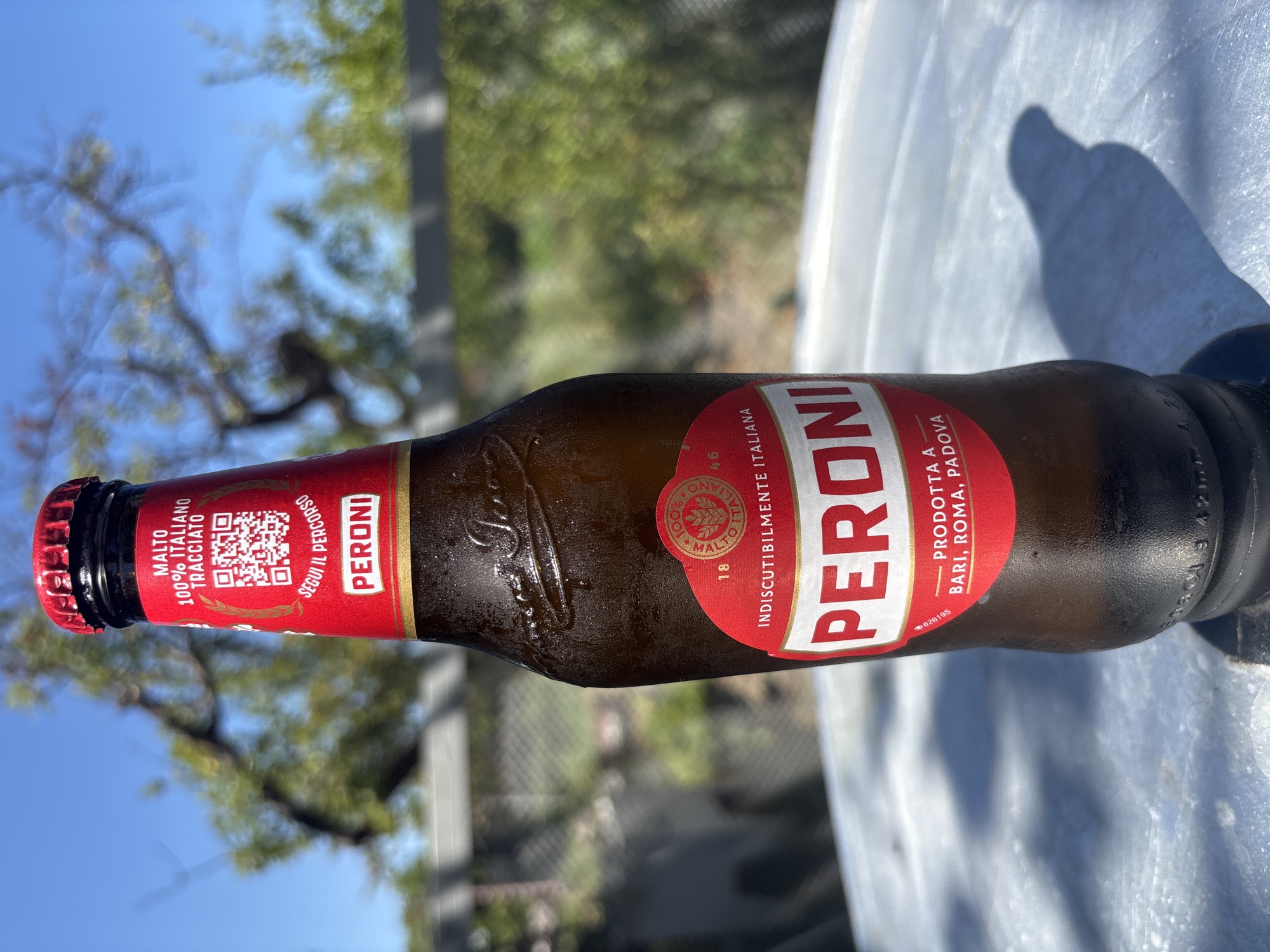A Peroni beer bottle is displayed outdoors, against a blurred background of trees and a blue sky. The bottle is condensation-covered and sits on a light-colored, textured surface. The label is predominantly red and white, featuring the Peroni logo, Italian text, and a QR code. The shadow of the bottle is cast onto the surface below.