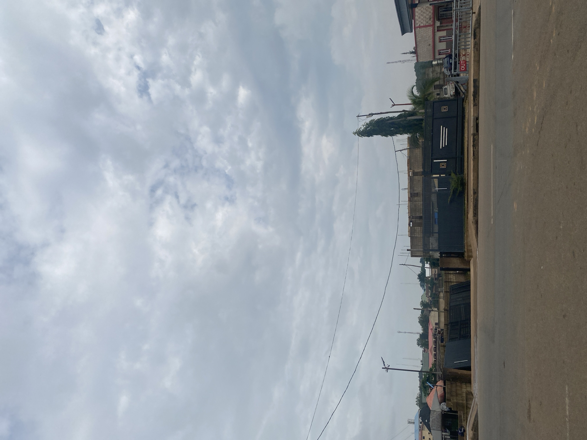 The image depicts an overcast sky over a street scene in Jos, Nigeria. A paved road occupies the foreground, with a sidewalk and a large, dark metal gate with a decorative pattern forming the mid-ground. Behind the gate, various buildings are visible, including structures under construction, a building with a red and white facade, and other residential and commercial buildings. Some trees, including a tall, slender one, dot the landscape. Power lines are strung across the sky, and utility poles are present. A few vehicles are parked near the gate and in the background. The overall impression is a typical urban environment on a cloudy day.