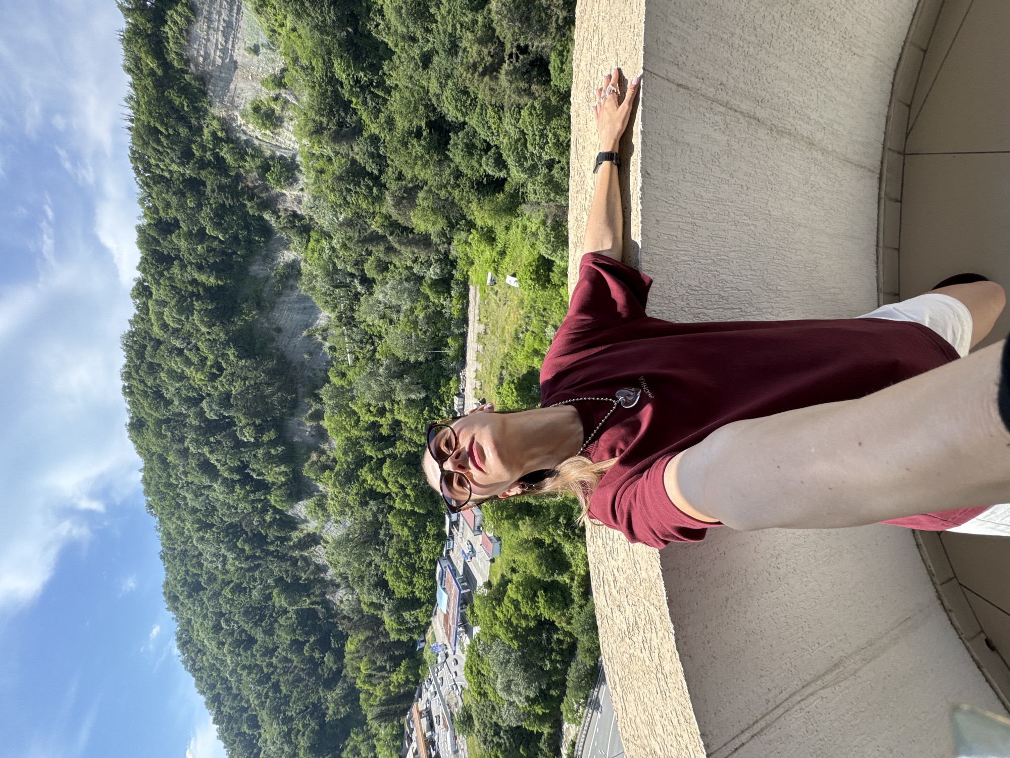 A woman stands on a balcony overlooking a dense, tree-covered mountainside in Sochi, Russia. The woman is wearing a maroon t-shirt, white shorts, sunglasses, and a necklace with a heart pendant. She is taking a selfie with her right arm extended, holding the camera. Her left arm is resting on the balcony railing, and she is looking upwards. In the background, a city street with cars and buildings is visible at the base of the mountain. The sky is blue with scattered white clouds. The balcony railing is made of textured concrete.