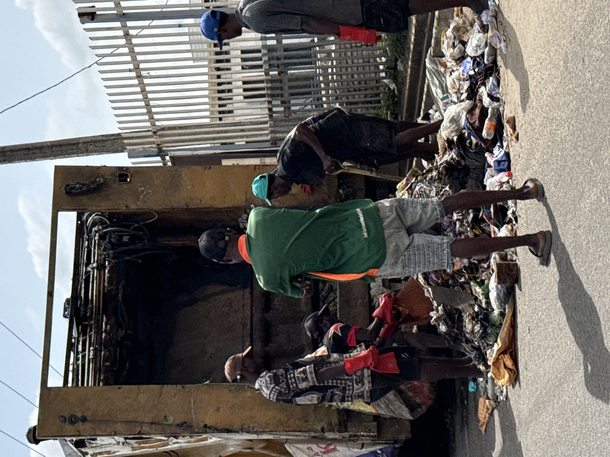 The image captures an outdoor scene in Ado, Nigeria, depicting individuals engaged in waste management activities. At least five men are visible, appearing to be collecting, sorting, or loading trash into a large, yellow, open-topped container, likely the back of a garbage truck or a skip.
In the mid-ground, a man wearing a green t-shirt with an orange trim, light shorts, and a black cap, equipped with red gloves, stands next to the large container. Another man, in a patterned shirt and a light cap, also with red gloves, is lower to the ground, interacting with the refuse. Further back, a man in a light blue cap and a dark t-shirt is bent over, seemingly inspecting or handling trash. To the left, a man in a blue cap and grey t-shirt, wearing red gloves, stands upright near a metallic slatted fence.
Piles of assorted refuse, including plastic bottles, bags, and fabric, are scattered on the asphalt or concrete ground around the container, indicating active waste handling. The background features a tall metal fence or railing under a clear, bright sky with some visible clouds.
Strong shadows cast on the ground suggest it is daytime, possibly mid-morning or afternoon, under direct sunlight. The scene portrays manual labor involved in urban waste collection or recycling efforts. No discernible text is clearly legible on clothing or items within the trash.