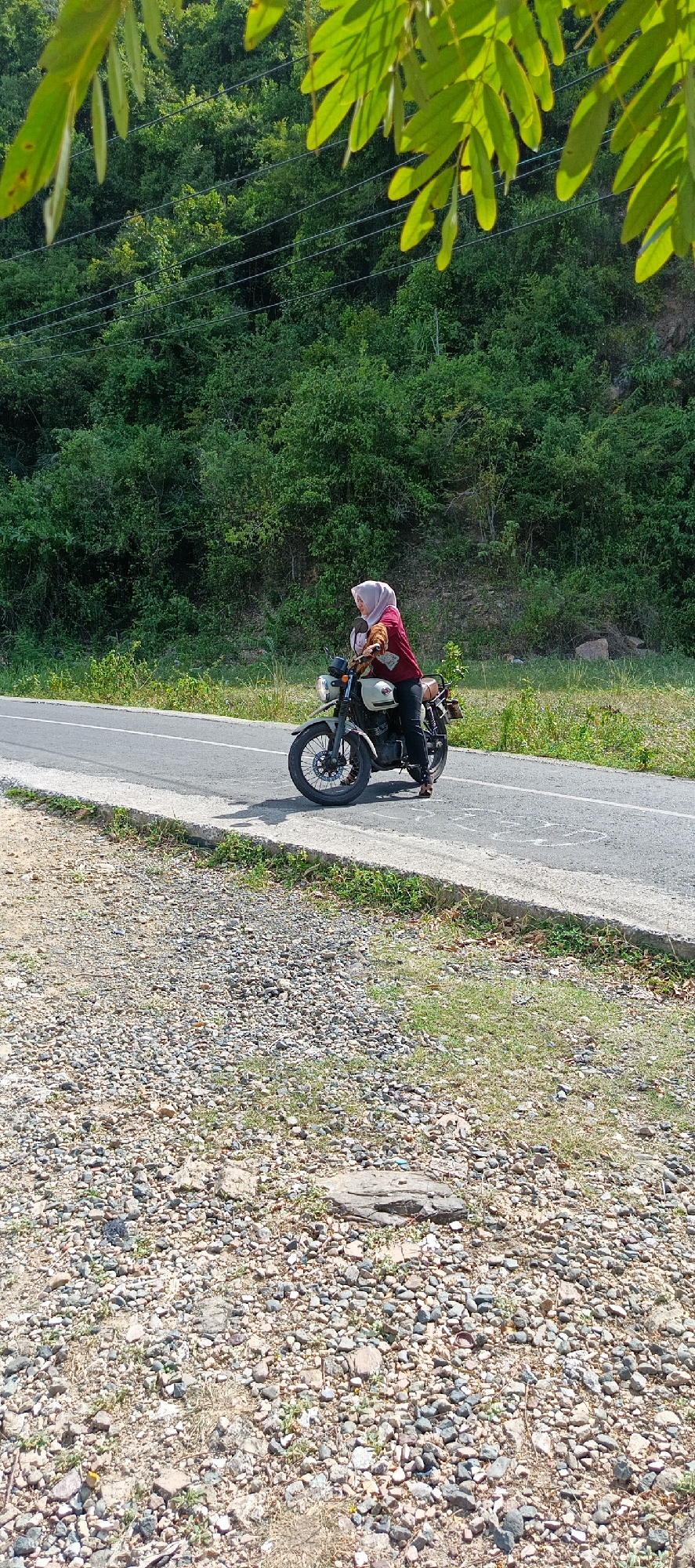 In Leuue, Indonesia, a person wearing a light pink hijab and a maroon shirt is seated on a cream-colored motorcycle. The motorcycle is positioned on the side of an asphalt road, with its front wheel turned slightly to the left. The road is bordered by a gravelly shoulder on the left and green vegetation on the right. In the background, a dense forest covers a hillside. The scene is illuminated by daylight, casting a shadow from the motorcycle onto the road.