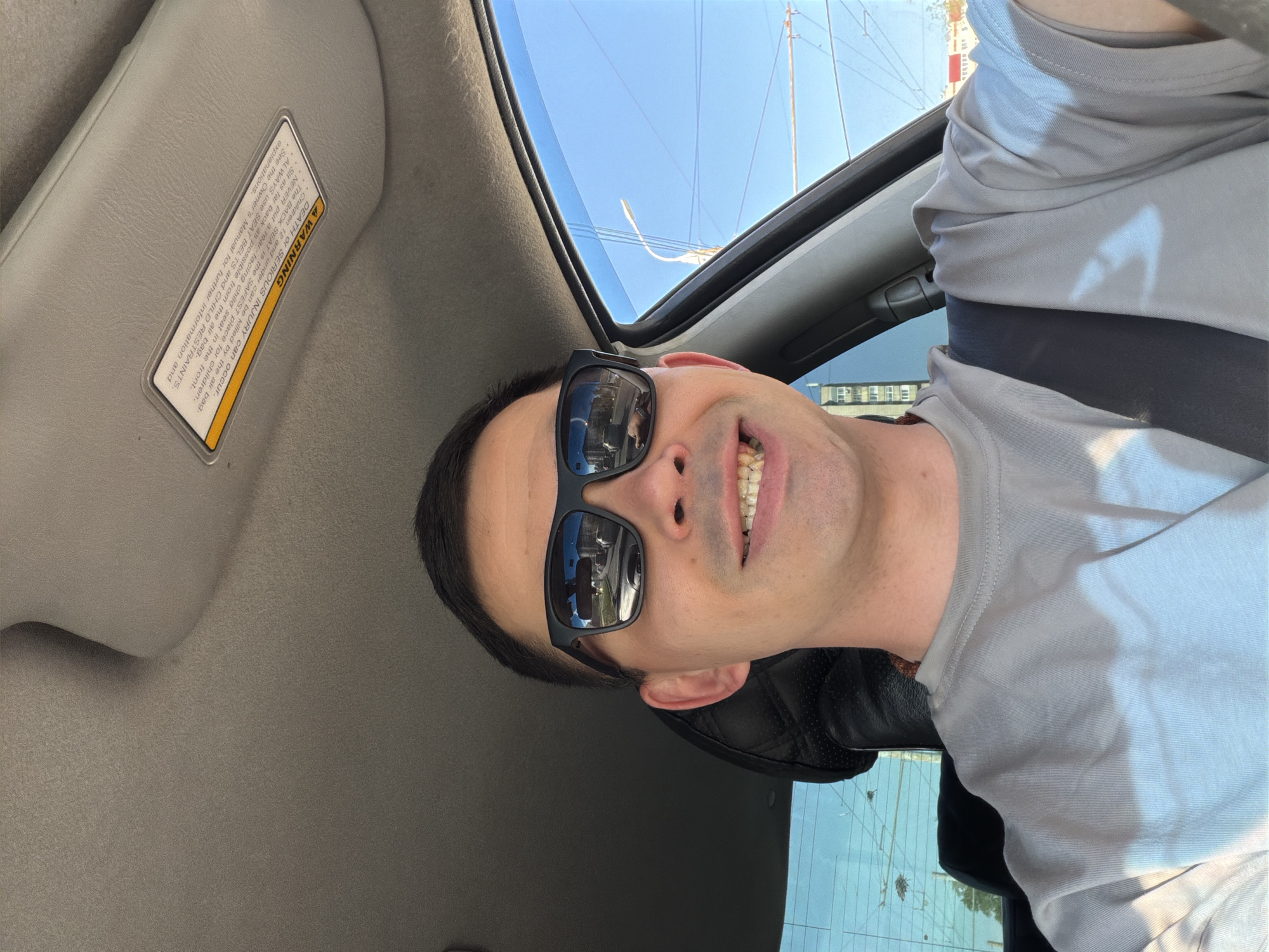 The image shows a man wearing sunglasses and a grey shirt sitting in a car with the sunroof open. The interior of the car is visible, including the sun visor with a warning label. The location appears to be Izhevsk, Russia, as the reflection in the sunglasses shows a vehicle with a license plate resembling a Russian license plate. The environment is sunny. The man is wearing a seatbelt.
