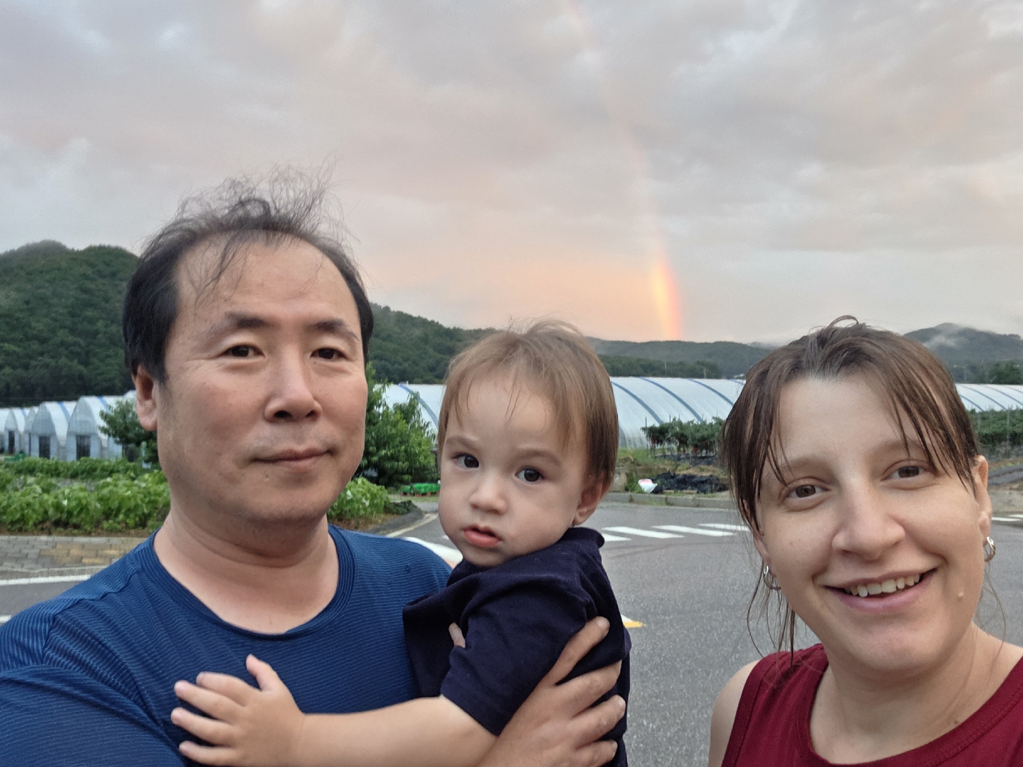 In an outdoor selfie, three individuals are captured against a backdrop of a cloudy sky with a faint rainbow. A man on the left, wearing a blue t-shirt, holds a young child in his arms. The child, dressed in a dark blue shirt, looks towards the camera. To the right, a woman with brown hair and a red tank top smiles at the camera. In the background, there are greenhouses and lush green hills. The scene appears to be set in a rural or semi-rural area, possibly the outskirts of Dunnae-myeon, South Korea, given the presence of agricultural structures like greenhouses. The overall atmosphere is casual and familial.