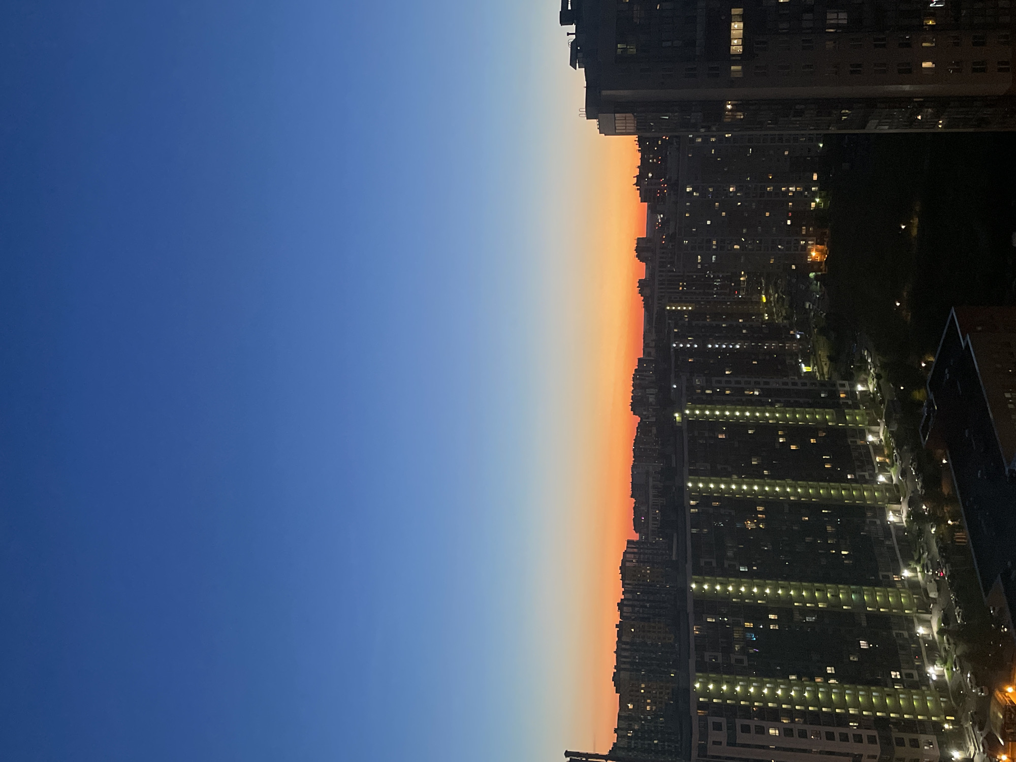 The image captures a dusk scene over an urban landscape in an unspecified location in Russia. The sky transitions from a deep blue at the top to vibrant orange and red hues near the horizon. Numerous multi-story residential buildings dominate the view, with many windows illuminated, indicating occupancy. In the foreground on the right, a tall, modern building with visible window lights stands prominently. To the left, a row of tall apartment buildings with vertical lighting strips is visible, featuring parked cars and some green spaces at their bases. The overall impression is one of a densely populated urban environment as evening sets in.