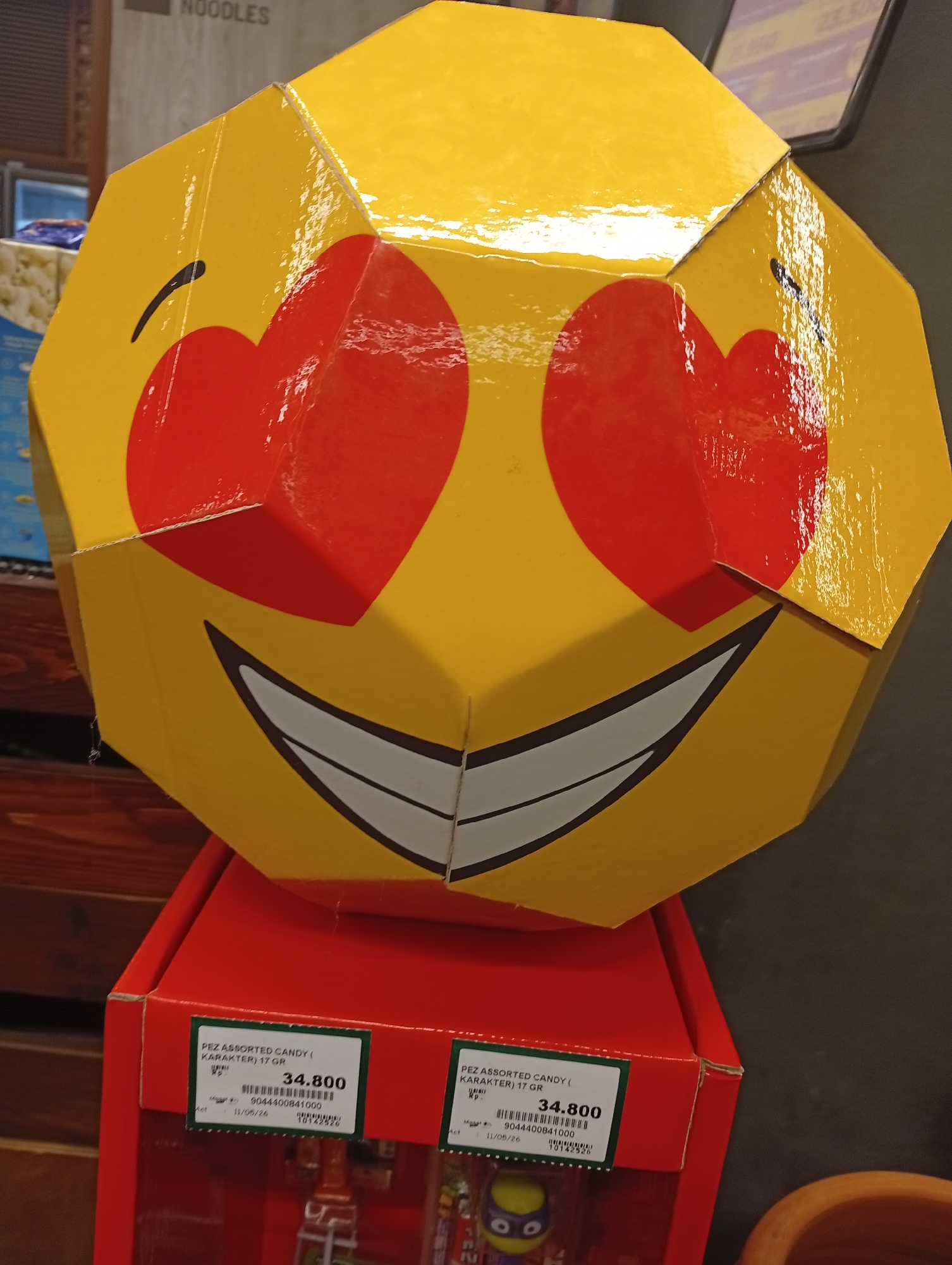The image shows a large, three-dimensional, yellow emoji with heart-shaped eyes and a wide, grinning mouth. The emoji appears to be made of assembled cardboard or similar material. It sits atop a red box labeled "PEZ ASSORTED CANDY (KARAKTER) 17 GR", with a price tag of 34,800 (presumably Indonesian Rupiah). The label includes the number "9044400841000" and the date "11/06/26". The photo seems to have been taken indoors in a commercial environment, likely a shop or store, in Denpasar, Indonesia, as indicated by the price and label information. Partially visible in the background are shelves and signs for other products, including "NOODLES".