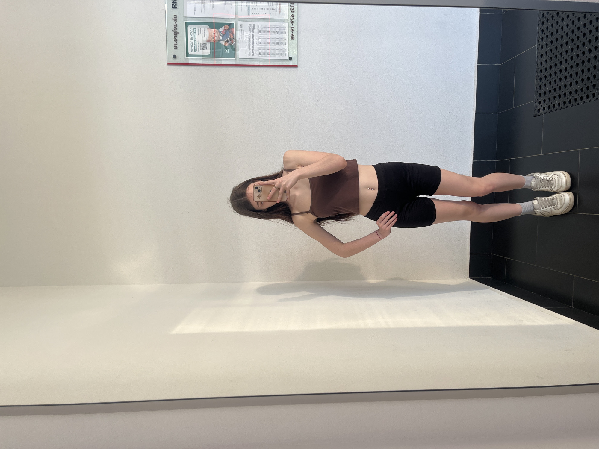 The image depicts a young woman taking a full-length mirror selfie in an indoor setting within an unknown city in Russia. The woman, with long, dark brown hair, covers her face with a light-colored smartphone featuring a patterned case. She wears a brown spaghetti-strap crop top, exposing her navel piercing, and black form-fitting shorts. Her footwear consists of white crew socks and off-white Nike Air Force 1-style sneakers. A thin red string is visible on her left wrist. She poses with her left hand on her hip, while her right hand holds the phone.
The environment features plain white walls, a black tiled floor with a black baseboard, and a black textured rubber mat on the floor to the right. A prominent rectangular information board with a red frame is mounted on the right wall. The board displays Cyrillic text, which appears mirrored in the reflection, along with a QR code and an image of a cat in a wheelchair. Visible reversed text elements include "ru.orglos-un.", what appears to be a Russian phone number "82-81-970 (21)", and other Cyrillic phrases related to instructions or organizational information. A distinct shadow pattern, consistent with window blinds, is cast on the left wall from an external light source.
