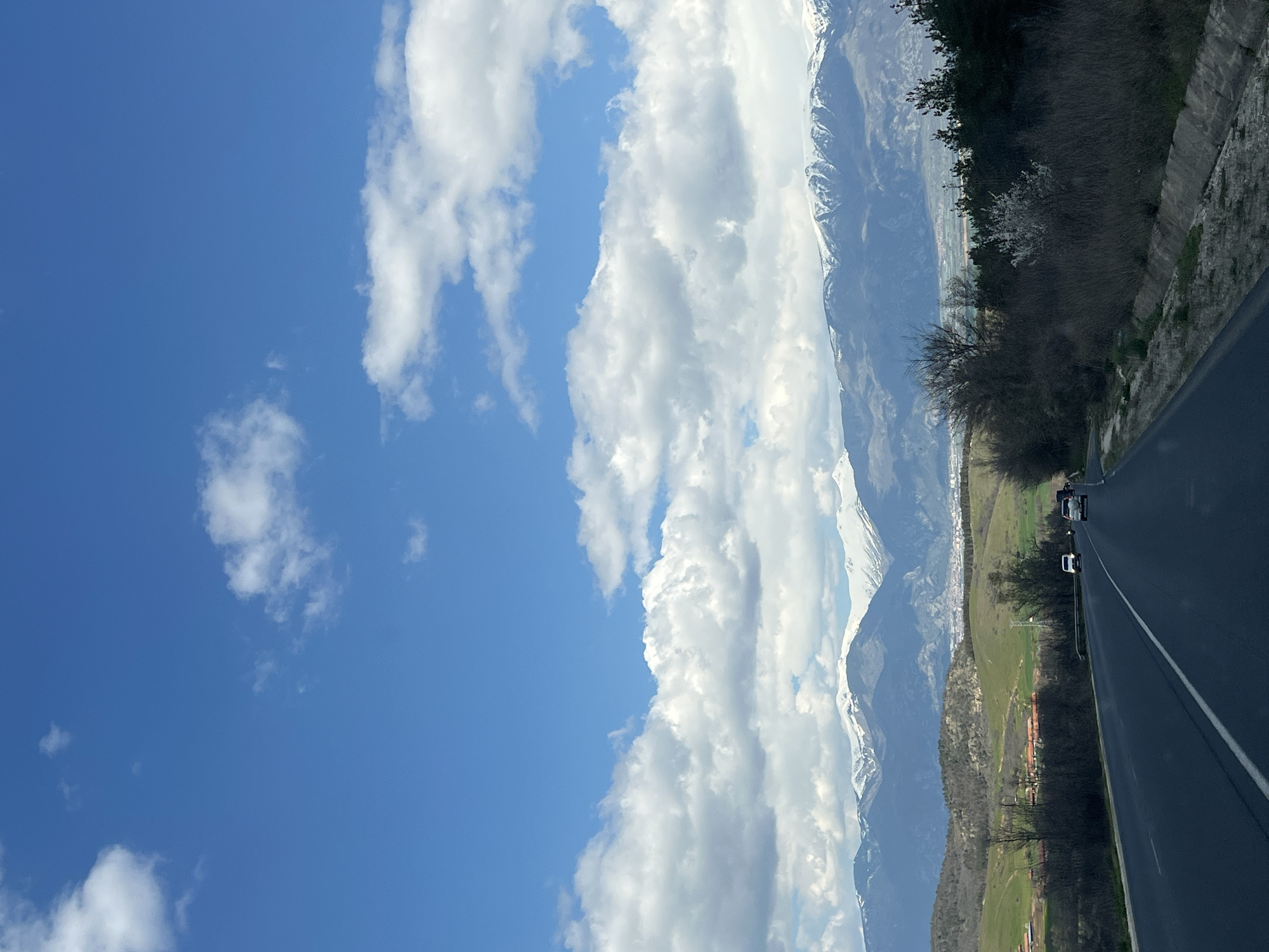 The image shows a view from a road near Mihiltsi, Bulgaria. To the left is a bright blue sky with scattered white cumulus clouds. Adjacent to the sky are mountains with snow-capped peaks. A road runs from the bottom right corner towards the center, with a white line dividing the lanes. Two vehicles are visible on the road. The area between the road and mountains appears to consist of fields and some scattered trees and vegetation. The terrain is generally green.