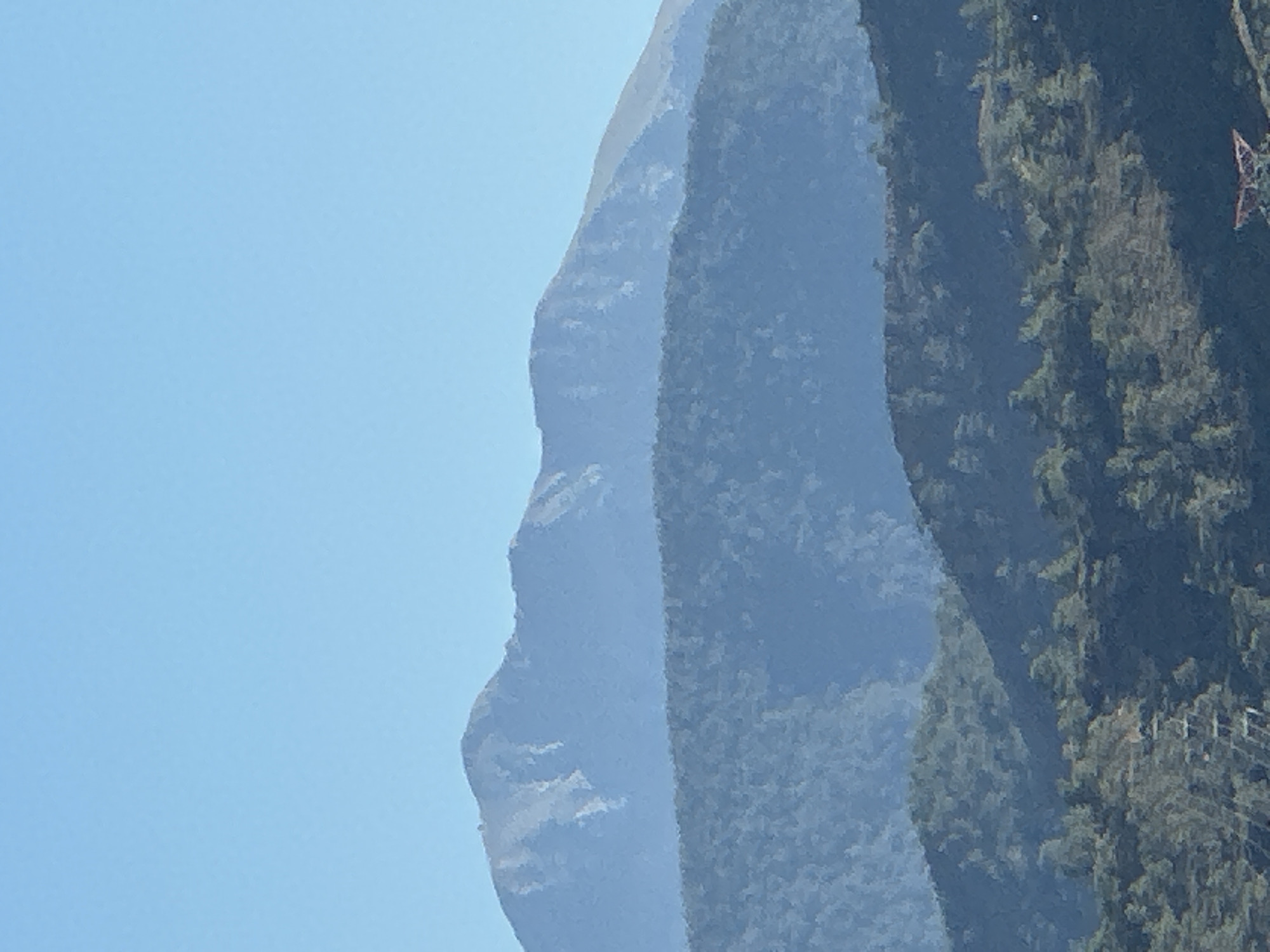 The image shows a landscape with mountains in Fethiye, Turkey. The sky is a clear blue. The mountains are arranged in layers, with the closest one covered in dense, dark green vegetation. A powerline structure is visible on the right side of the closest mountain. The mountains behind it are lighter in color and appear more distant, with the furthest being hazy blue.