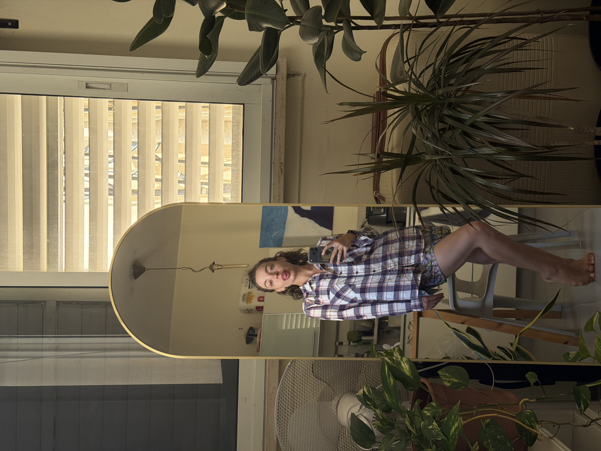 A young woman is captured in a mirror selfie, standing barefoot in what appears to be an apartment in Tel-Aviv, Israel. She is wearing a plaid button-down shirt and patterned shorts, with her tongue playfully sticking out towards the camera. The background reveals a window with horizontal blinds, a ceiling light fixture, a fan, several potted plants, and furniture, suggesting a domestic setting. The lighting in the room is warm and diffused, likely from the natural light coming through the window.