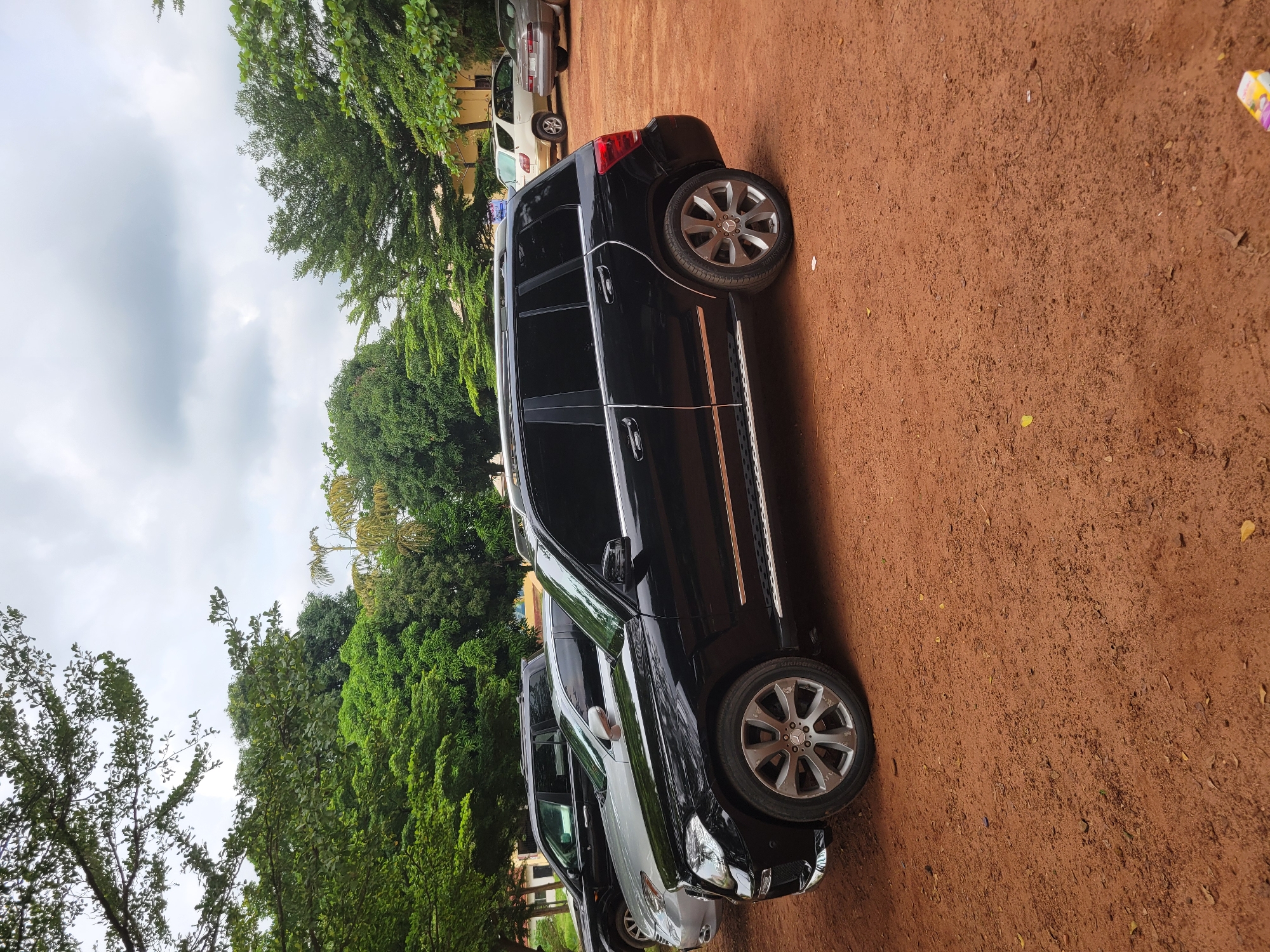 The image shows a black Mercedes-Benz SUV parked on a dirt road in Ukukwa, Nigeria. The car is the primary subject, with its side profile prominently displayed. The vehicle has tinted windows and chrome accents. To the left of the SUV, another lighter-colored SUV is partially visible. In the background, lush green trees and vegetation are abundant, suggesting a natural or semi-rural setting. The sky is overcast, indicating a cloudy day, possibly with the threat of rain. The ground is covered in reddish-brown dirt, typical of many areas in Nigeria. There are no people visible in the image, and the focus is solely on the parked vehicles and the surrounding environment. The scene appears to be an outdoor setting where vehicles are parked, possibly a roadside or a private lot. The time of day is not clearly discernible due to the cloudy weather, but it appears to be daytime. There is no visible text in the image.