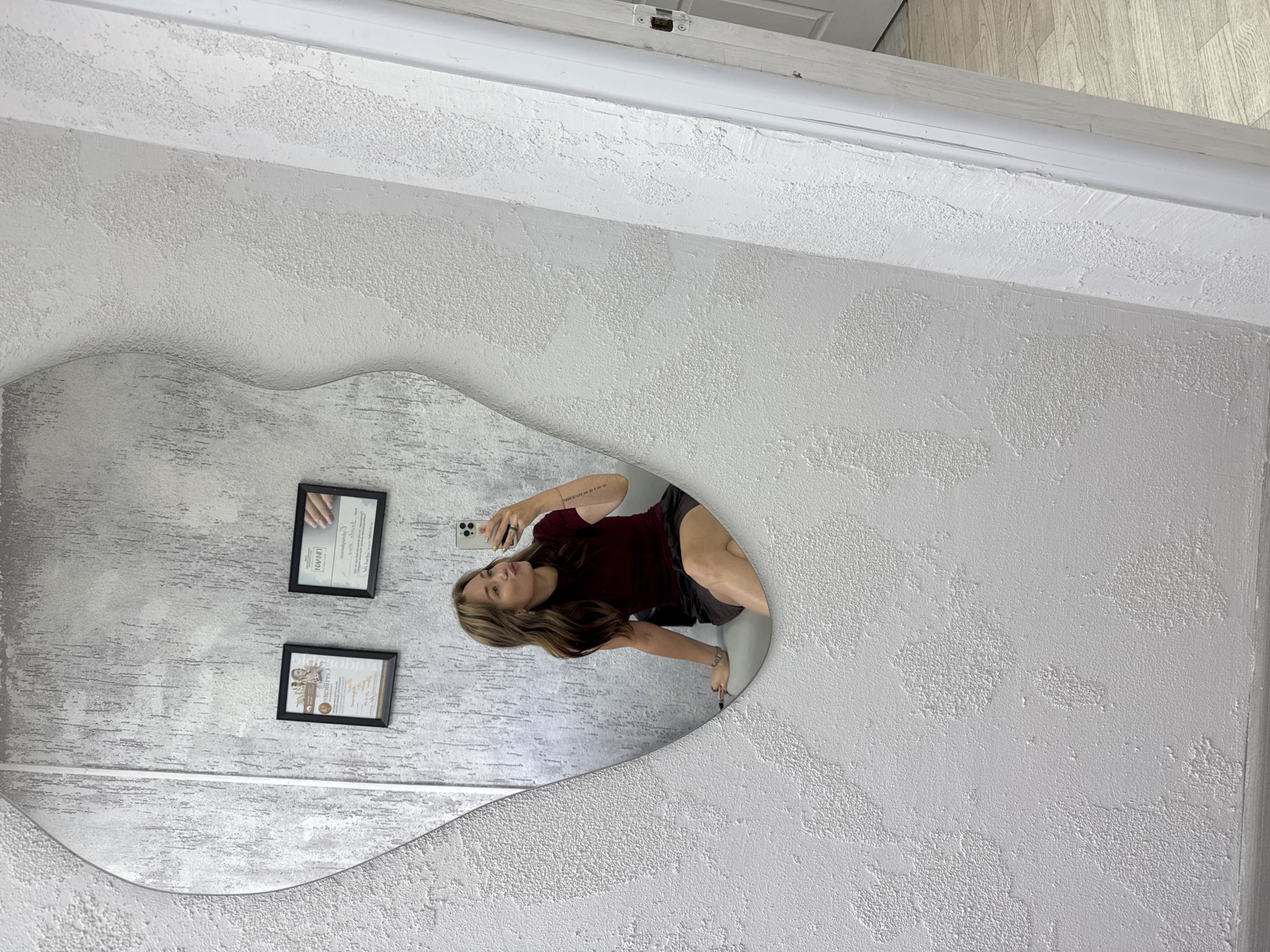 In an indoor, brightly lit environment, a young woman with brown hair, wearing a burgundy top and dark shorts, sits on a grey bench. She is taking a selfie in a large, irregularly shaped mirror that reflects her image. The mirror is mounted on a textured, light grey wall. To the left of the woman in the reflection, two framed certificates are hanging on the wall. The floor appears to be light wood. To the right of the mirror, a white door frame is visible, suggesting an entryway or exit. The overall aesthetic is modern and clean, with a focus on texture and reflective surfaces. The specific location in the city of Chaikovsky, Russia, is not discernible from the image.