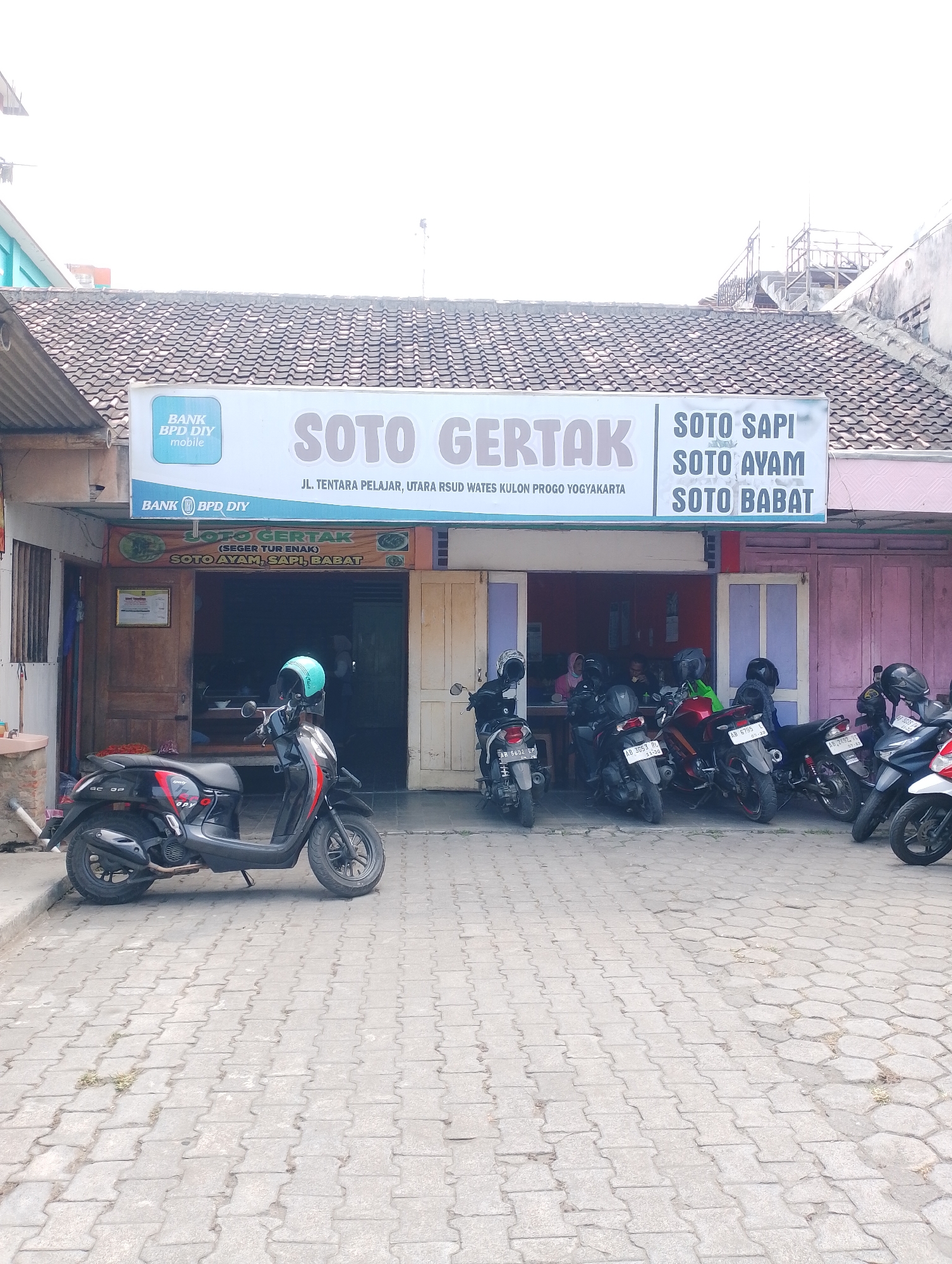 The image depicts the exterior of a restaurant called "Soto GERTTAK" in Wates, Indonesia. The restaurant is situated along a street, indicated by the presence of parked motorcycles. The building features a tiled roof and a facade with multiple doorways and windows. A prominent sign above the entrance advertises "SOTO GERTTAK" and lists menu items such as "SOTO SAPI," "SOTO AYAM," and "SOTO BABAT." Another smaller sign indicates the address as "JL. TENTARA PELAJAR, UTARA RSUD WATES KULON PROGO YOGYAKARTA." Several motorcycles are parked in front of the establishment, suggesting it is a popular local eatery. Inside, glimpses of patrons can be seen seated at tables. The foreground is paved with interlocking brick pavers. The scene is illuminated by daylight, with a bright sky suggesting an outdoor setting.