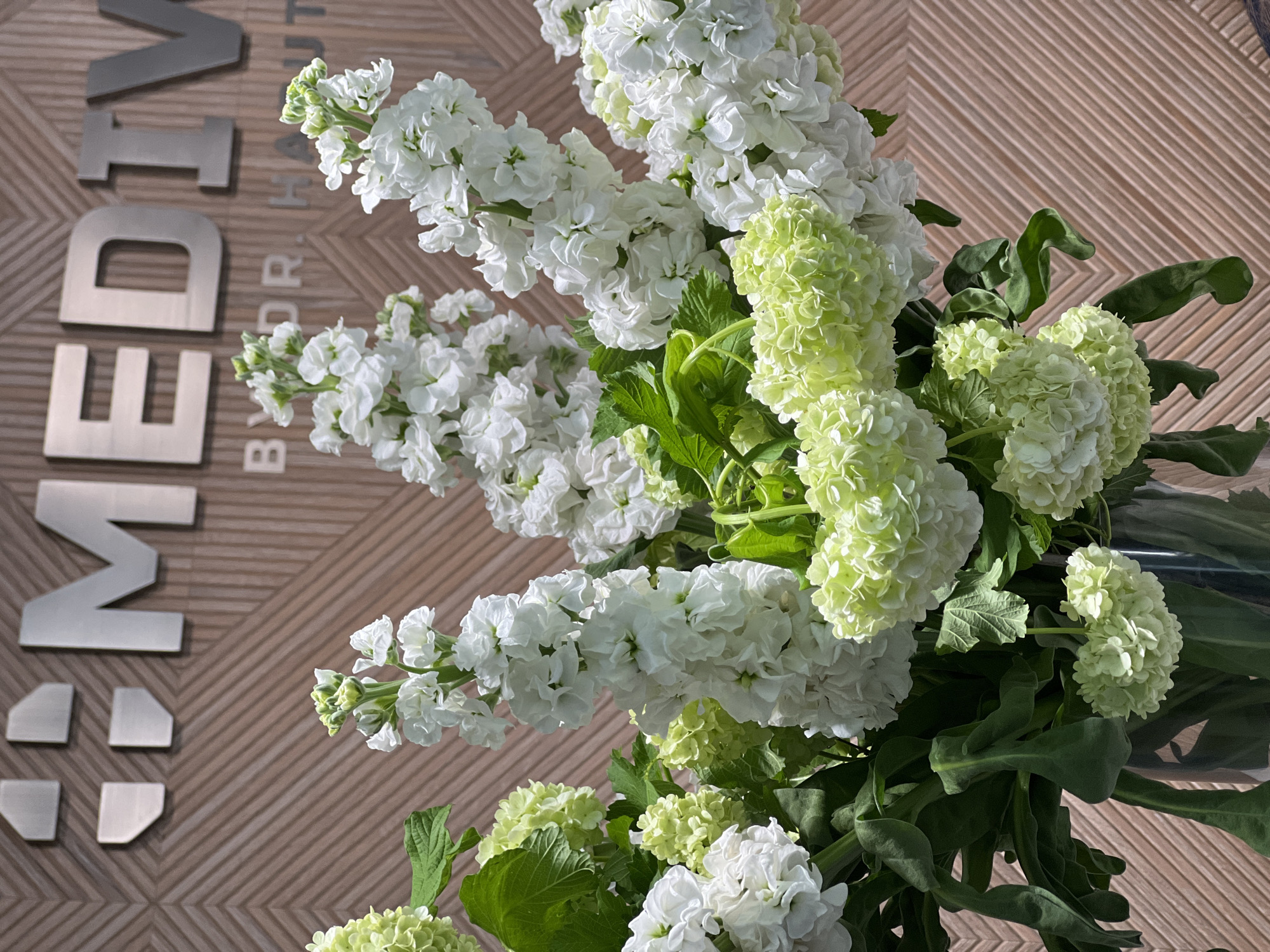 A high-angle shot features an arrangement of white and light green flowers, potentially stocks and hydrangeas, placed in front of a wall with metal letters reading "MEDIA BY DR." The wall has a geometric pattern of light brown wood. The arrangement is positioned towards the right side of the image, while the letters are on the left. Based on the letters, the image could be taken in or near the "Media by Dr." office. The exact location within Yerevan, Armenia cannot be determined from the visual data provided.