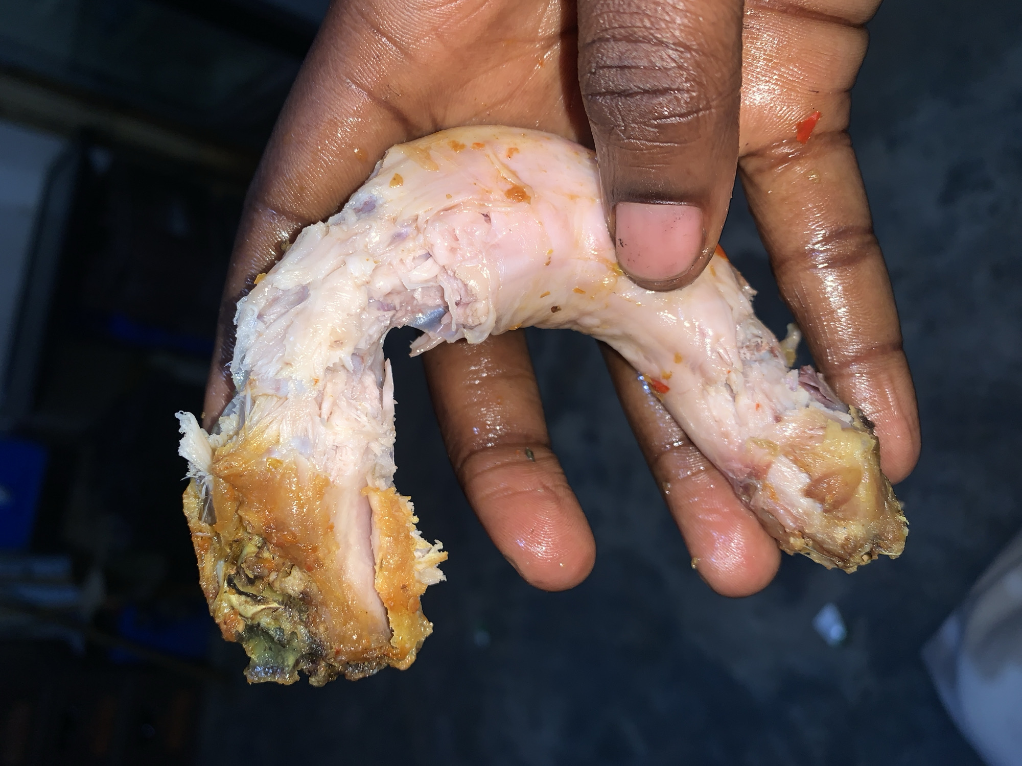 A dark-skinned hand holds a cooked, partially consumed chicken neck. The hand appears oily or wet. The chicken neck shows visible bone and cooked meat. The surrounding environment is dark and out of focus. There is no indication of the location being New Stadium, Nigeria.