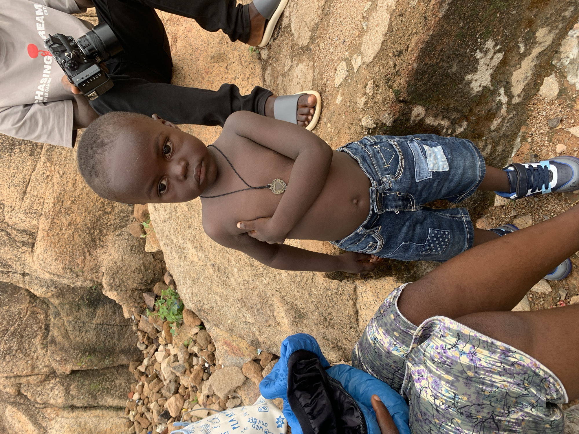 A young, shirtless, dark-skinned boy wearing denim shorts and blue sneakers stands on a light brown rock formation. He has a pendant necklace and is looking at the camera with his arms crossed. An adult sits beside him, and a person with a camera is visible in the upper left corner. The location is likely in a rocky area of Gura Top, Nigeria.
