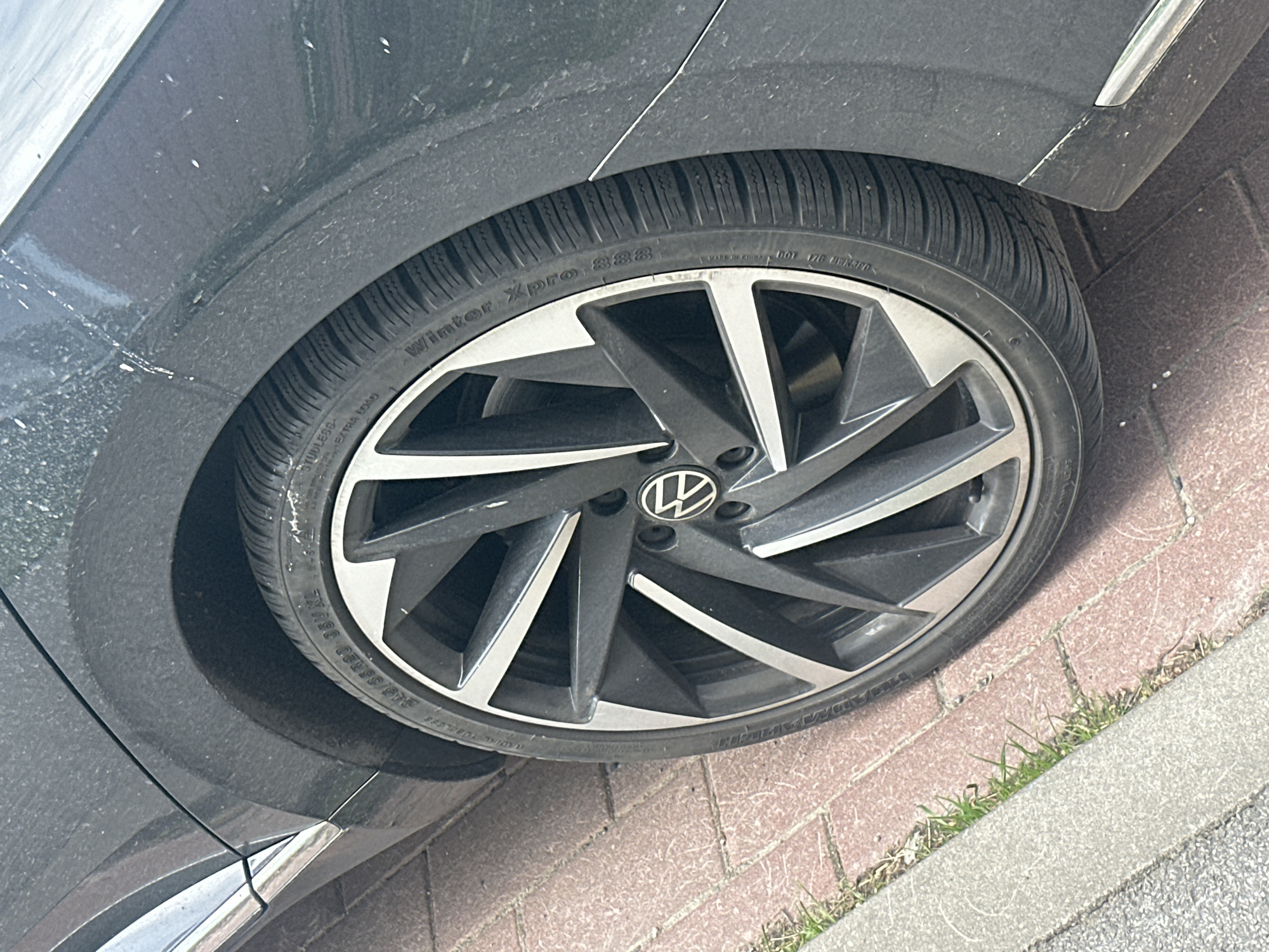 The image shows a close-up of a dark-colored car wheel and tire, parked on a brick-paved street. The tire is a "Winter Xpro 388" model with a size indication of "215/55R20 95V XL". The wheel is a multi-spoke alloy design with silver and dark grey accents. A Volkswagen logo is visible on the center cap of the wheel. The car's bodywork, also dark-colored, is visible in the background and appears dusty. The street is paved with red bricks, and a section of a concrete curb and some grass are visible to the left. The lighting suggests it is daytime. The image was captured in Tallinn, Estonia, as indicated by the metadata.