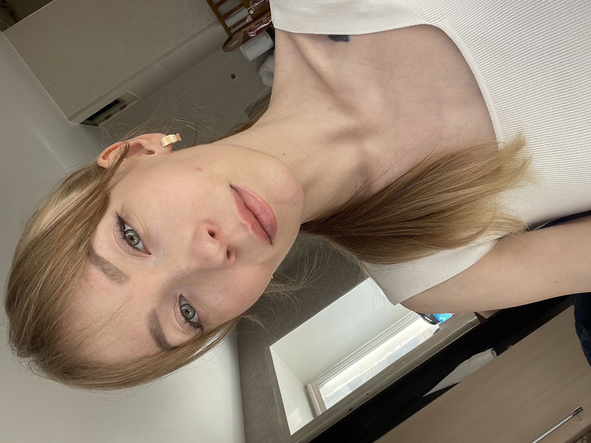 A close-up, tilted shot of a light-skinned woman with blonde hair and light-colored eyes. She is wearing a white, textured top. A gold-colored rectangular earring is visible on her left ear. There is a small black tattoo on her upper chest/neck area. The background includes a window with natural light coming through, a glimpse of a desk or table, and a section of a wall and ceiling. A partial view of a closet or storage unit is also visible.
There is no evidence to suggest this image was taken in Perm, Russia.