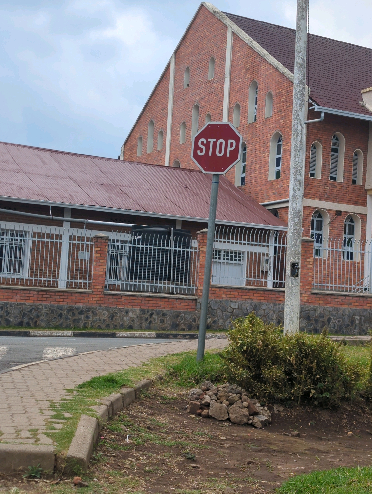 The image depicts a stop sign in front of a red brick building with a dark red roof in Musanze District, Rwanda. The building appears to be a church or a similar large institution, with arched windows and decorative brickwork. A white metal fence runs along the front of the building, with a black water tank visible behind the fence on the right side. A paved sidewalk runs along the left side of the image, with a grassy area and a pile of rocks in the foreground. The sky is overcast. There are no people visible in the image.