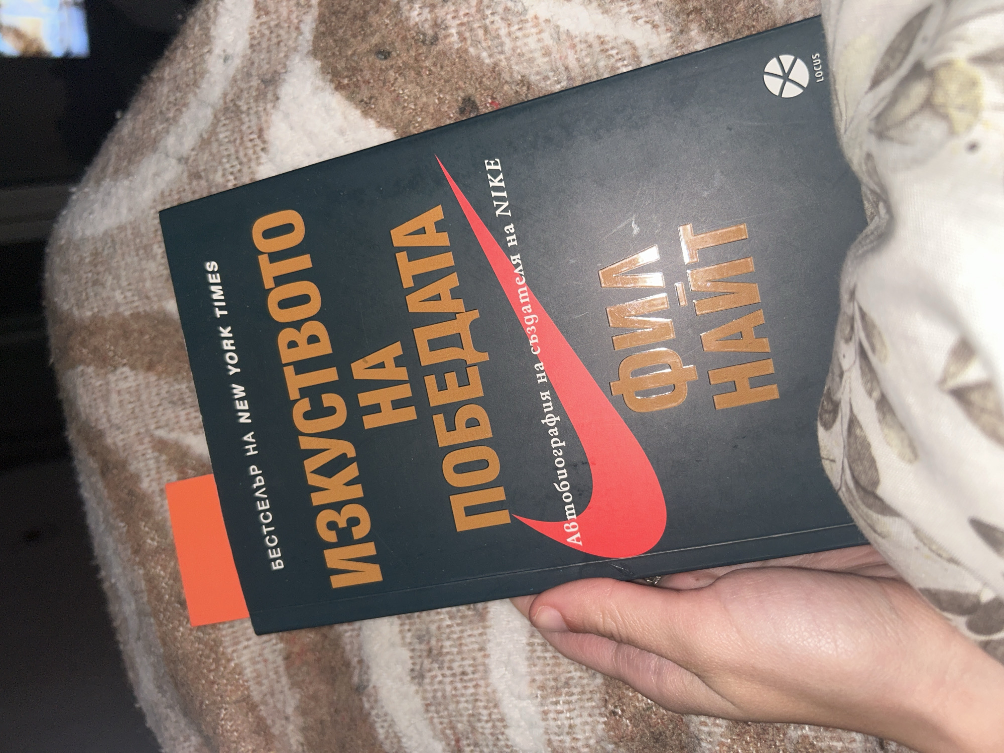 The image shows a hand holding a book titled "The Art of Victory" (ИЗКУСТВОТО НА ПОБЕДАТА), which is the autobiography of Nike founder Phil Knight. The book is a Bulgarian translation, as indicated by the Cyrillic script. The book cover is primarily dark green with gold lettering and a red Nike swoosh. A bookmark is visible on the left side of the book. The book appears to be resting on a patterned blanket or fabric. The location is unknown as the city is not specified in the image.