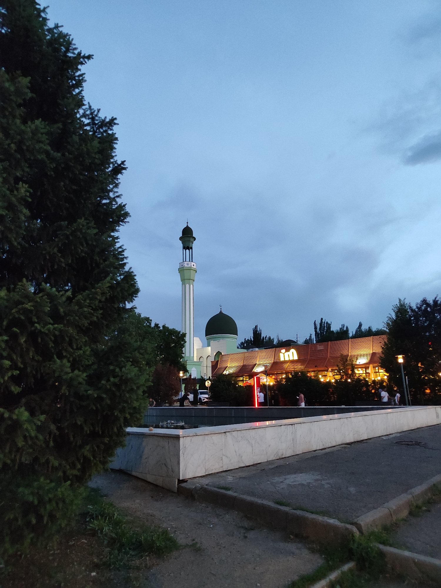 The image shows a view of a McDonald's restaurant and a mosque in Almaty, Kazakhstan, at dusk. In the foreground, a large evergreen tree is on the left, and a marble-edged fountain is in the center. The McDonald's building, illuminated with warm lights, is to the right of the mosque. The mosque features a tall minaret with a green dome and is adjacent to the restaurant. Several people are visible near the fountain and the McDonald's, some walking and others standing. The sky is overcast and a deep blue, indicating evening.