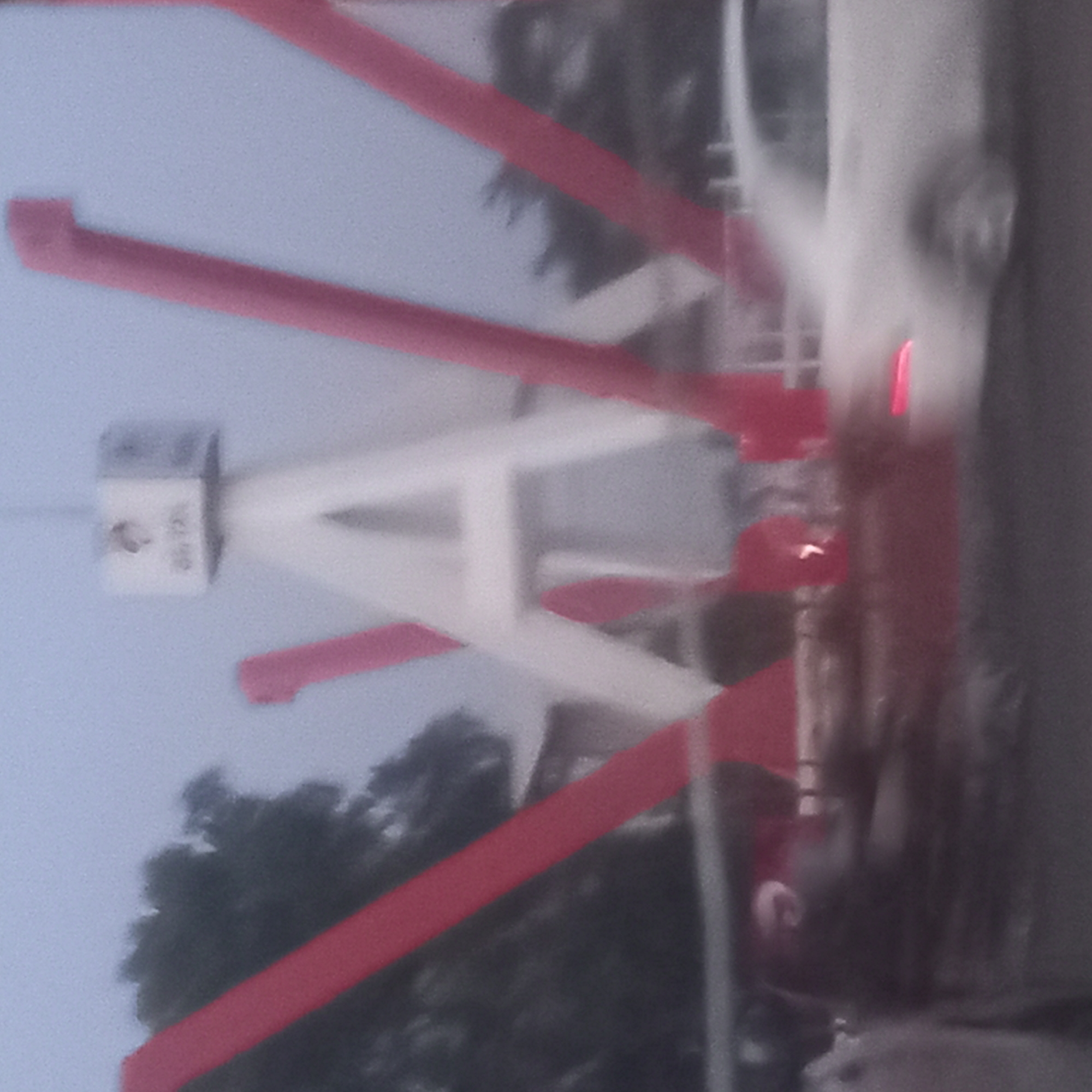 The image shows a blurred view of a white and red tower structure in Barnawa, Nigeria. The structure has a box-like top with text on it. The white structure has an A-frame with red supports that extend from the tower to the ground. Trees are visible in the background. The image appears to have been taken from a moving vehicle. A white car is visible in the background, partially out of frame.
