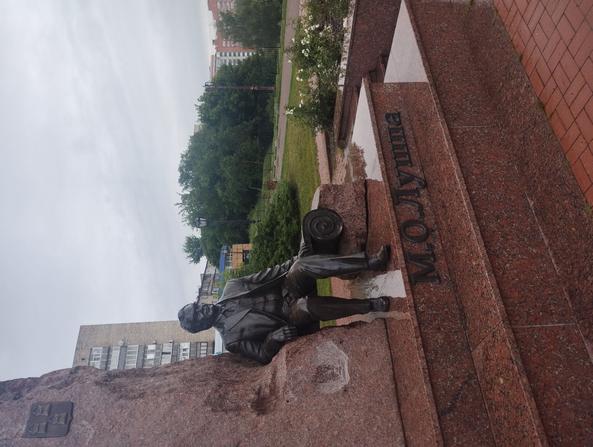 A bronze statue of a seated man in a suit and tie is positioned on a granite monument in Sumy, Ukraine. The statue is on the left side of the frame, leaning against a large, rough-textured red granite boulder. The man is holding a cylindrical object with a coiled pattern on its side. Below the statue, on a red granite step, is an inscription in dark letters that reads "M.O. LYUTSHA". The monument has multiple red granite steps leading up to it, some of which appear wet. In the background, there are trees, bushes with white flowers, a brick-paved walkway, streetlights, and apartment buildings. The sky is overcast. The overall scene suggests a public monument or memorial.