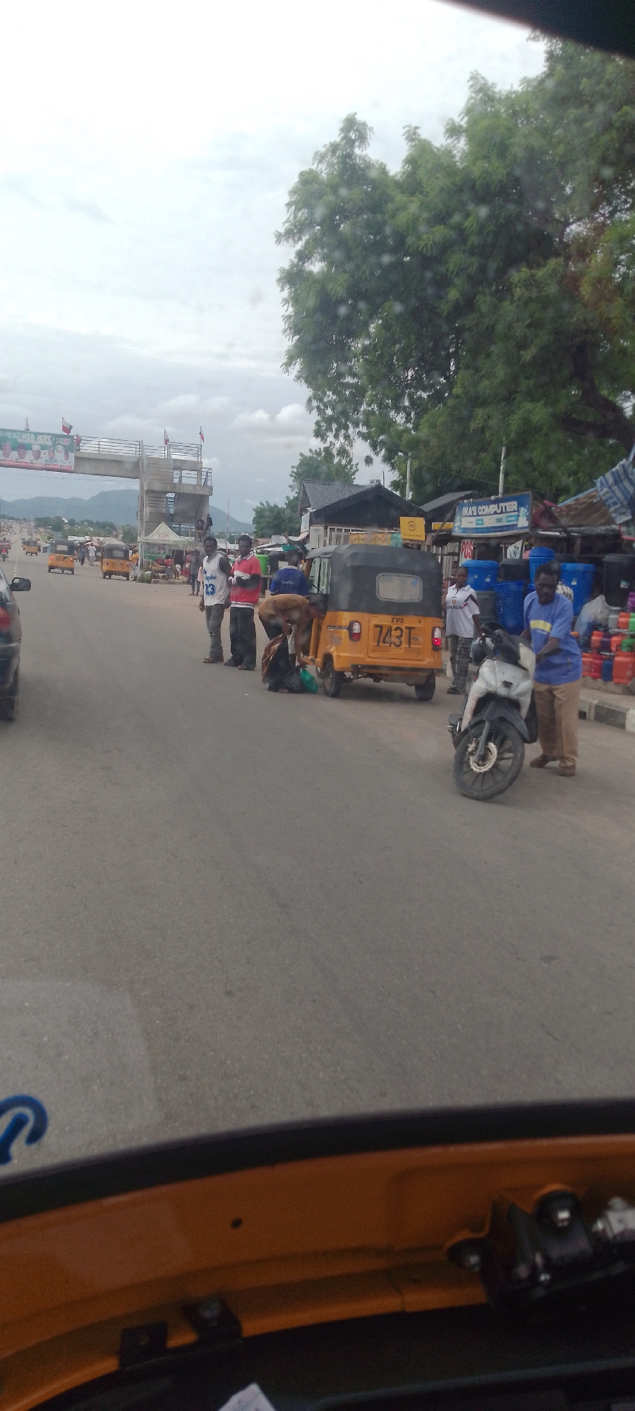 The image depicts a street scene in Jalingo, Nigeria, likely near a border crossing or a significant intersection, given the presence of an overhead archway with flags and banners. The road is paved and appears to be a main thoroughfare with several vehicles. In the foreground, from the perspective of someone inside an orange vehicle, the rear bumper and part of the interior of the vehicle are visible.
Further down the road, a yellow auto-rickshaw (also known as a Keke Napep) is parked, with its license plate reading "743T". Several individuals are gathered around the auto-rickshaw, some appearing to be loading or unloading items. One person, wearing a blue shirt and khaki pants, is standing to the right of the auto-rickshaw. Another person in a white shirt with "23" on it and dark pants is standing near the auto-rickshaw, looking towards it. To their left, a person in a red shirt and black pants is also present. A few people are crouched down near the back of the auto-rickshaw, engaged in activity.
To the right of the auto-rickshaw, a white motorcycle is parked, with a person in a blue shirt and khaki pants standing next to it. Behind the motorcycle, a shop front is visible with various items displayed, including what appear to be containers or barrels. A sign for "IKA'S COMPUTER" is prominently displayed above this shop. Further down the road, more auto-rickshaws and other vehicles are visible, along with buildings and trees. The sky is overcast, suggesting it might be a cloudy day. The overall impression is one of a busy, everyday street scene in a Nigerian urban environment.
