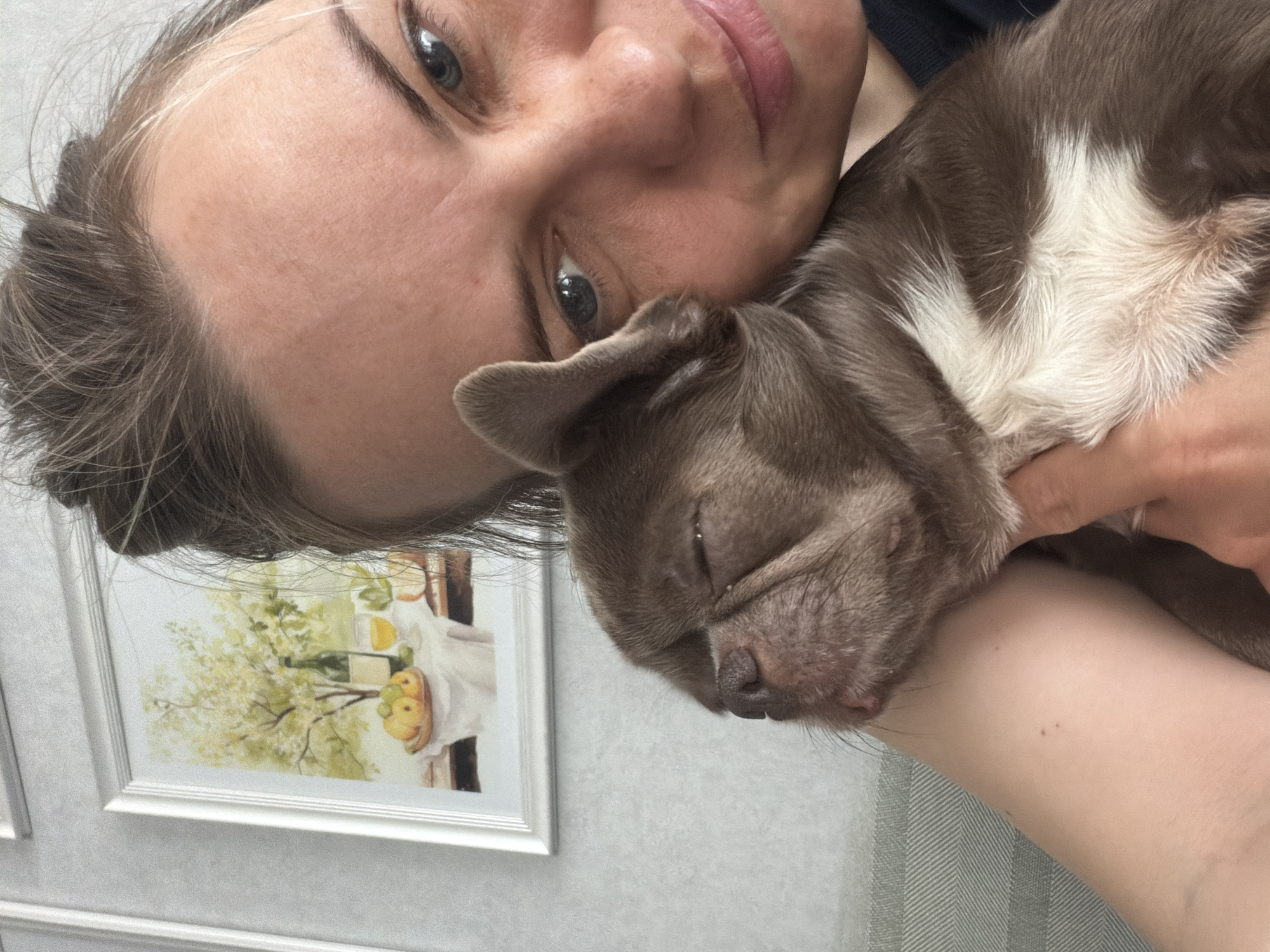A woman with fair skin and blue eyes is holding a brown and white puppy close to her face. The puppy appears to be sleeping or resting. In the background, a framed painting is visible on the wall. The location is Votkinsk, Russia.