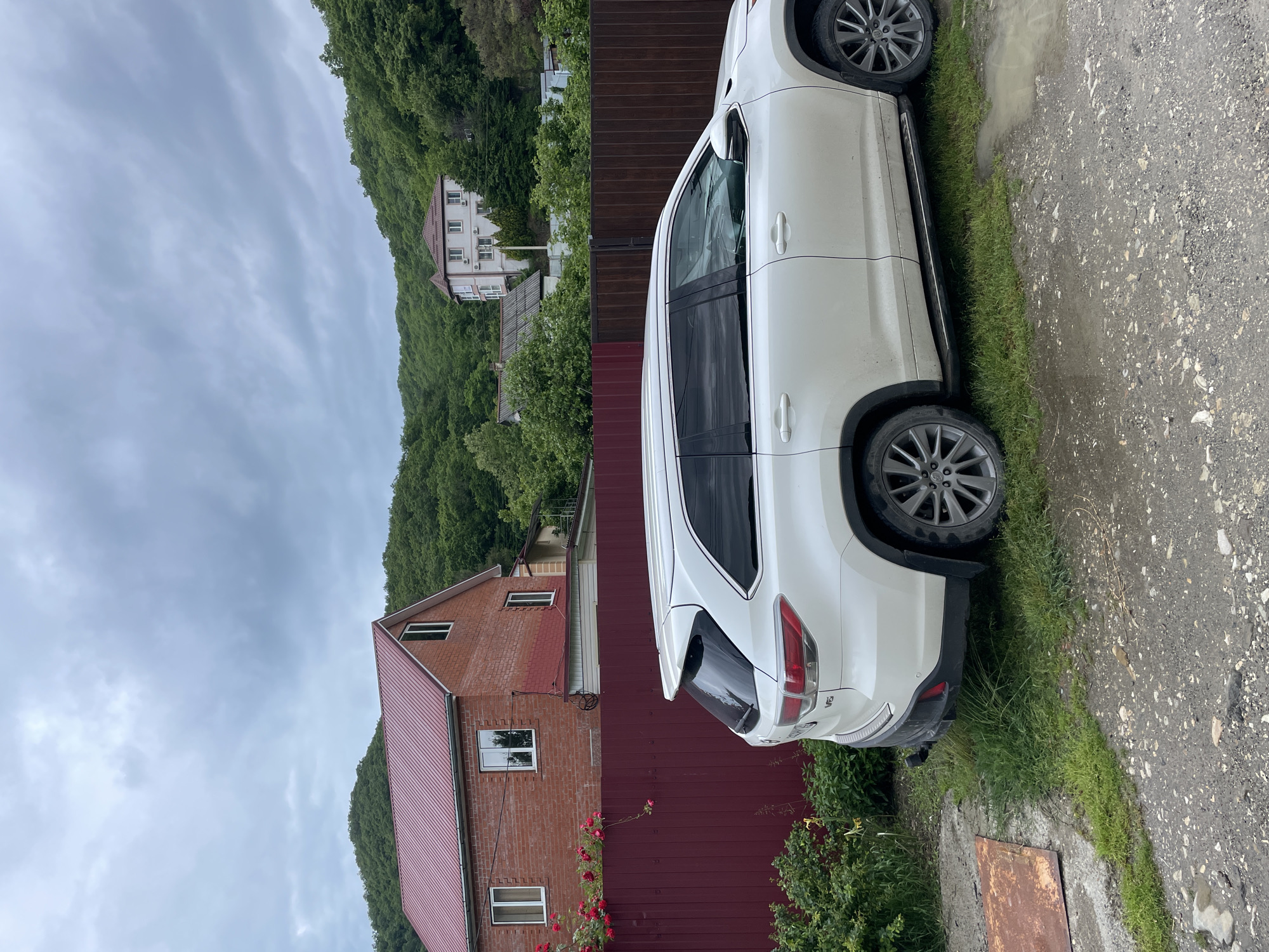 The image shows a white SUV parked on a patch of grass next to a gray concrete surface. A burgundy fence is visible behind the vehicle, with a brick building with a red roof adjacent to the fence. In the background, a hillside covered with trees is visible, along with a white building with a red roof. The sky is overcast. The SUV is likely a Toyota Highlander, judging by the visible features. The image suggests a residential area near a hillside. The city location is Olginka, Russia.